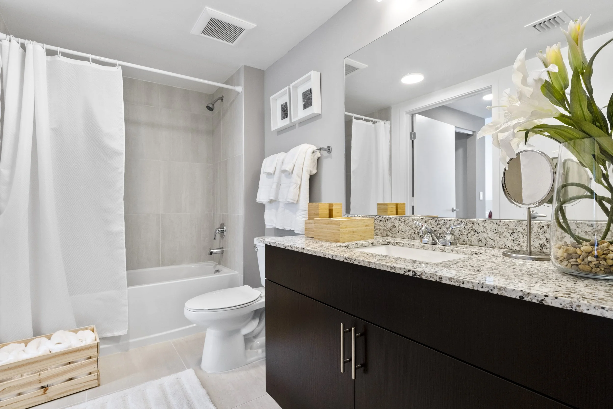 Modern bathroom with a bathtub and shower, white towels, a granite countertop with a sink, mirror, decorative flowers, and framed artwork, featuring a neutral color palette with grey walls and black cabinetry.
