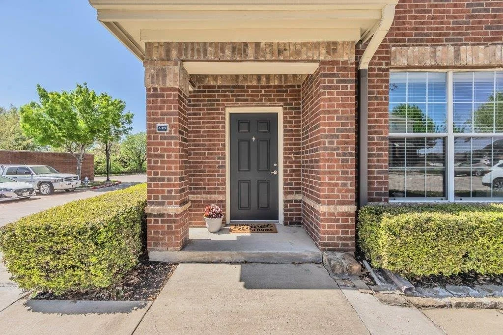 Front entrance of a brick apartment building with a black door, potted plant, and welcome mat. Surrounded by a concrete walkway, trimmed bushes, and parked cars in a parking lot.