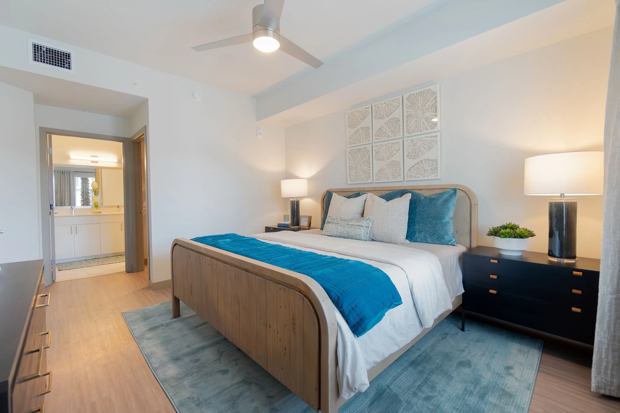 A modern hotel bedroom with a wooden bed frame, white bedding, blue throw blanket, and assorted pillows. Two black nightstands with lamps and decorative items flank the bed. A wall art piece is above the headboard, and a ceiling fan is overhead. An a