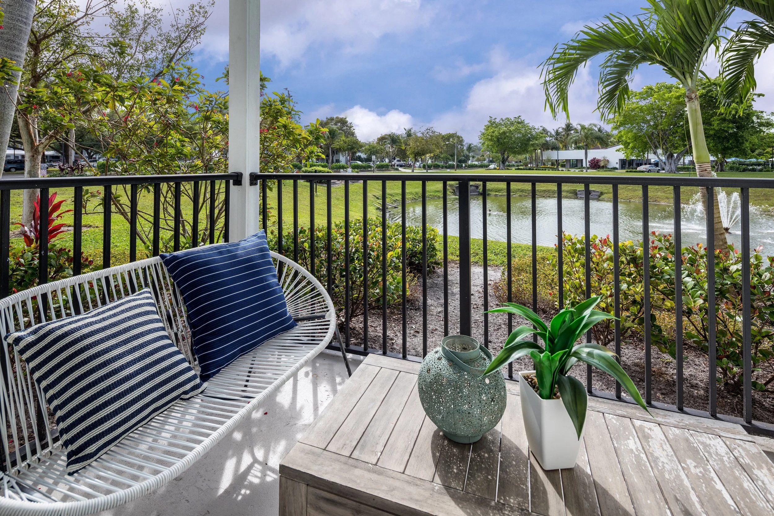 View from a patio with a white wicker bench holding two striped blue pillows, a potted green plant, a decorative vase, overlooking a pond with a fountain, lush green bushes, trees, and a partly cloudy sky.