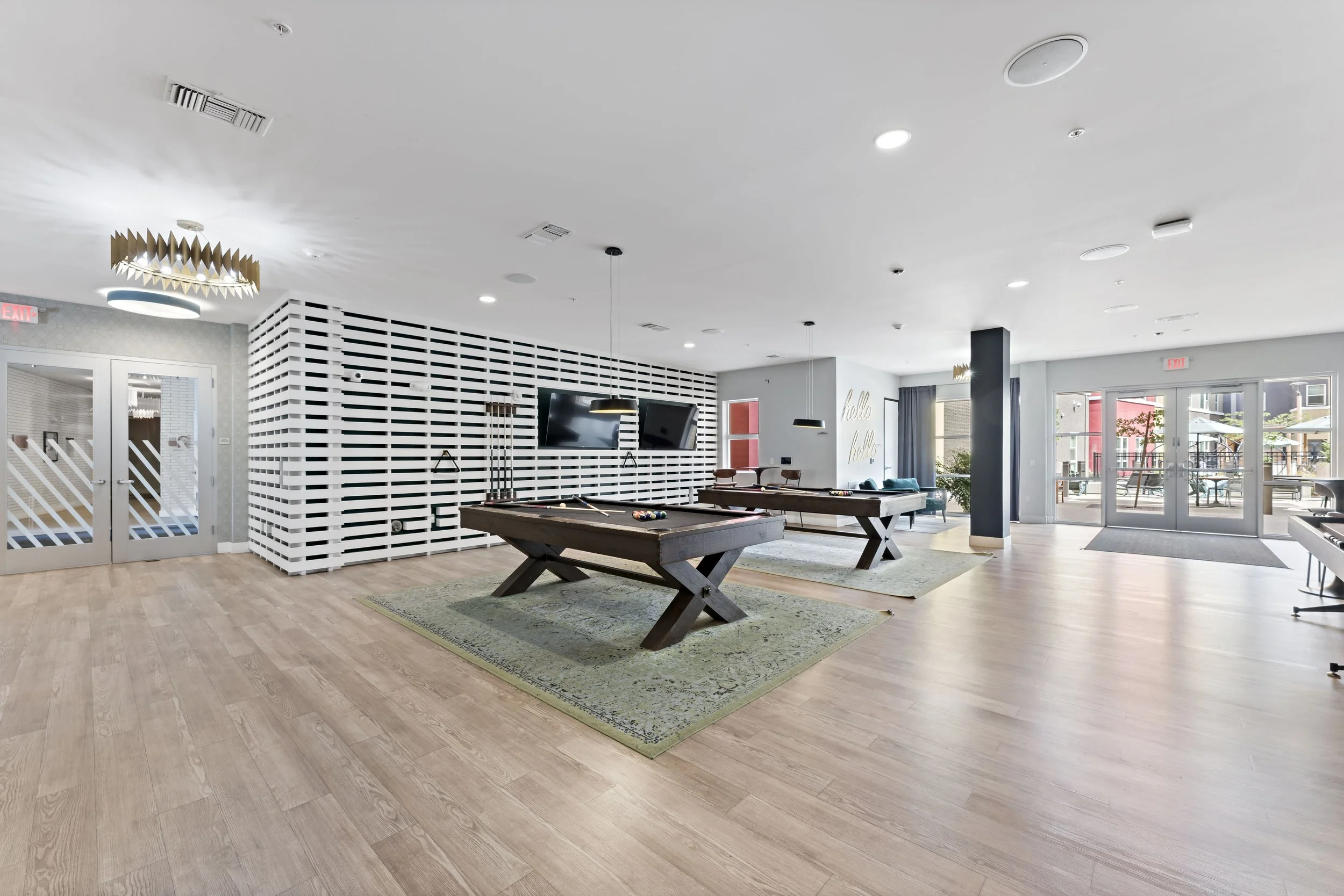 An interior space with two pool tables on green rugs, a wall with black and white horizontal stripes, multiple ceiling lights, a TV mounted on the wall, and a seating area with lounge chairs near large glass doors leading outside.