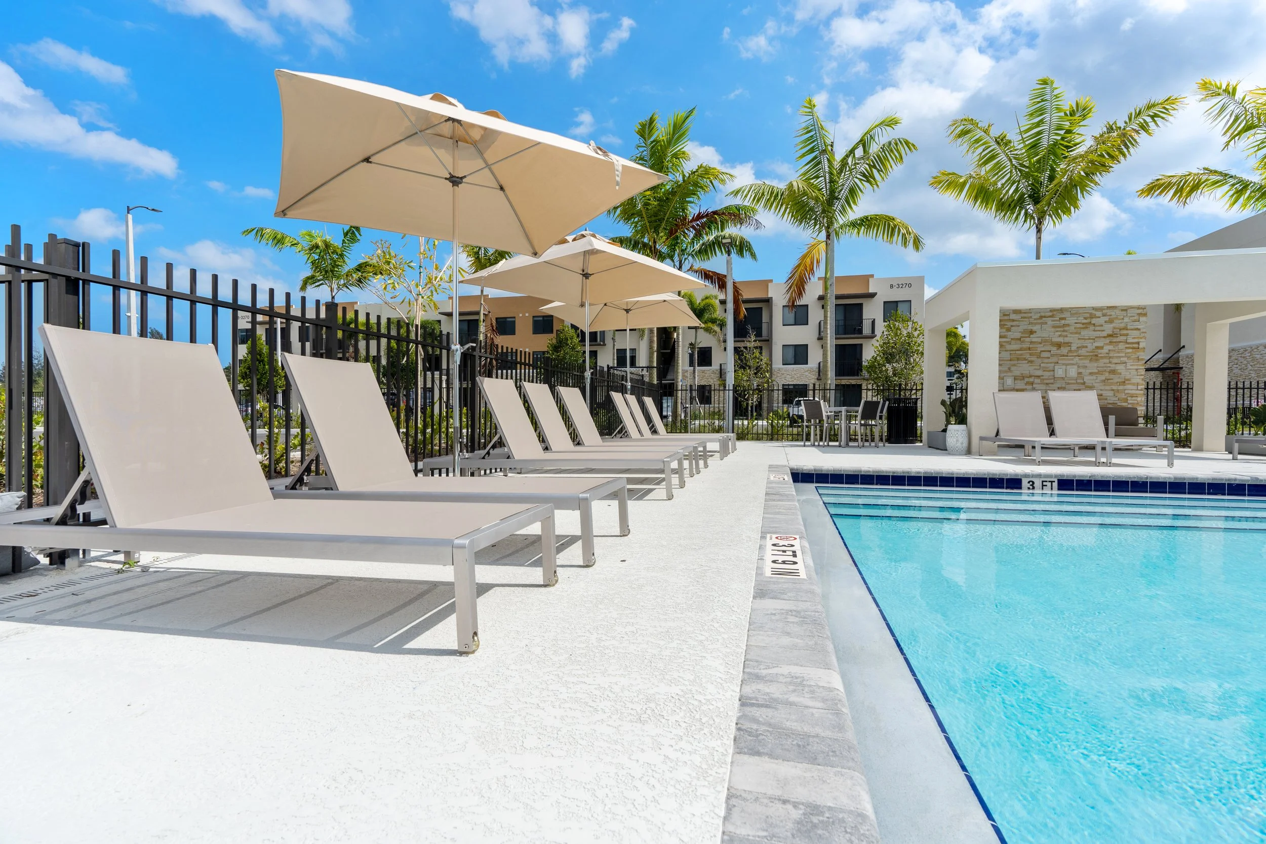 Poolside area with lounge chairs, umbrellas, and green palm trees under a partly cloudy blue sky.