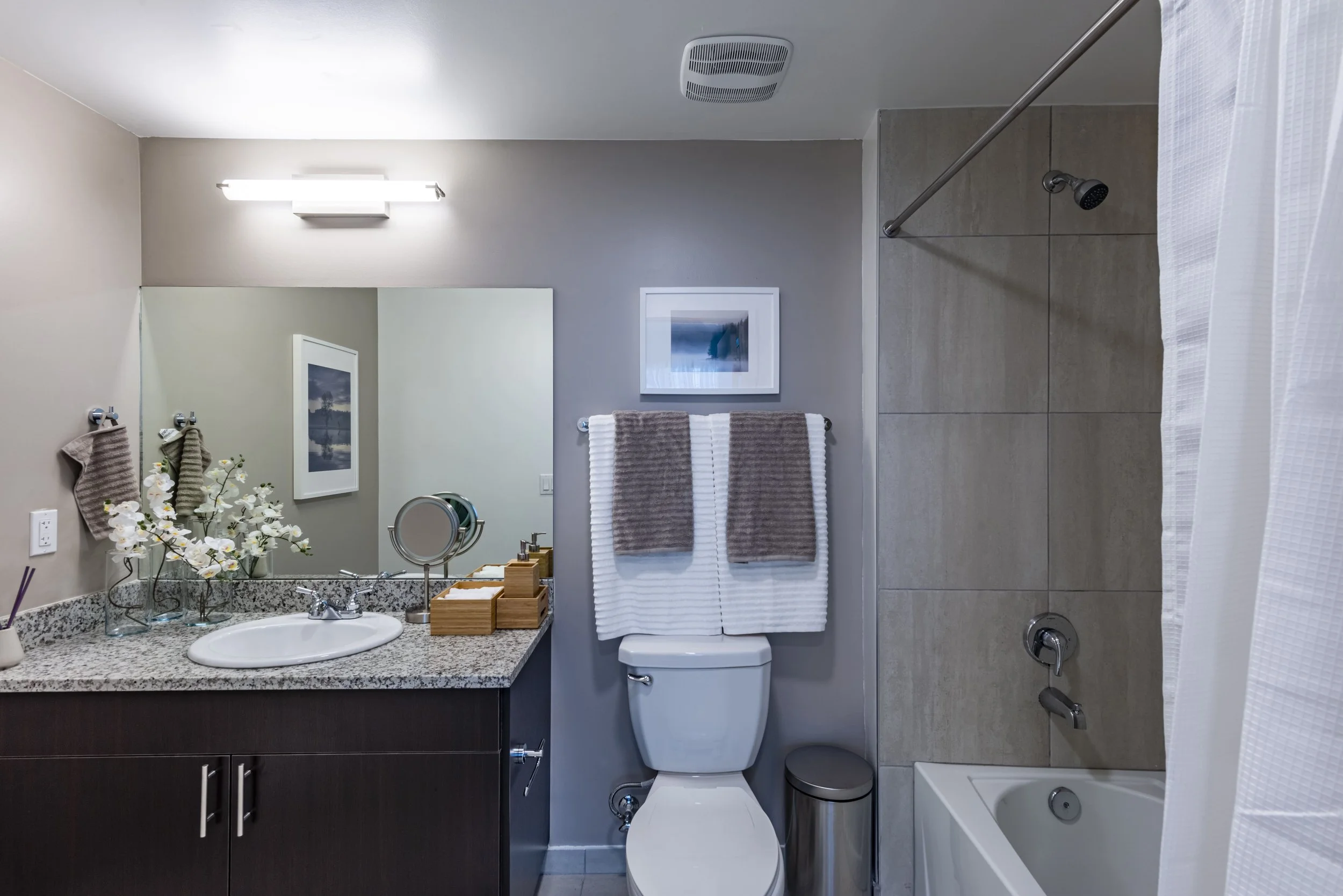 A modern bathroom with a large mirror, granite countertop, white sink, flowers, and a light fixture above. There are two grey and white towels hanging above the toilet, which is next to a shower with a bathtub, textured wall tiles, and a showerhead. 