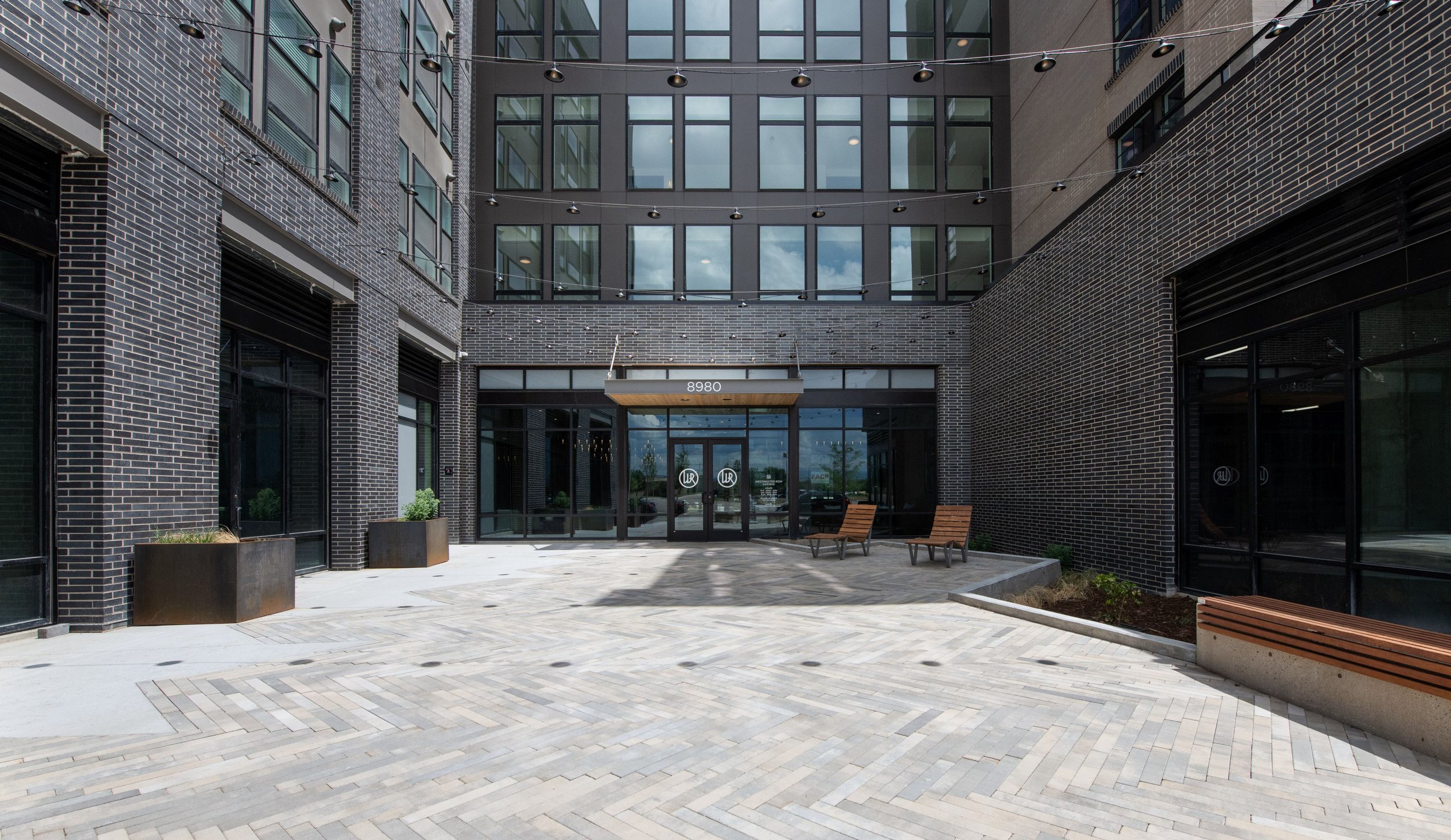 Modern apartment building entrance with glass doors, brick walls, two wooden benches, and potted plants in outdoor planters.