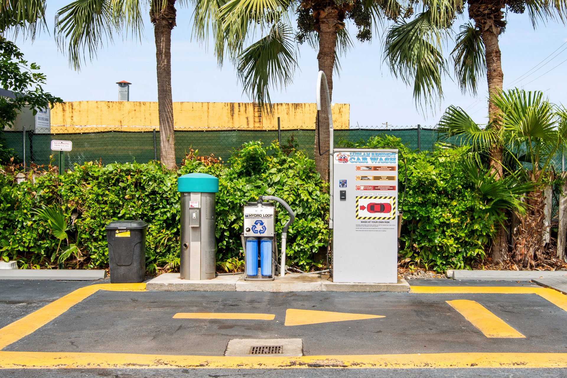 Self-service car wash station with parking space, surrounded by palm trees and greenery.