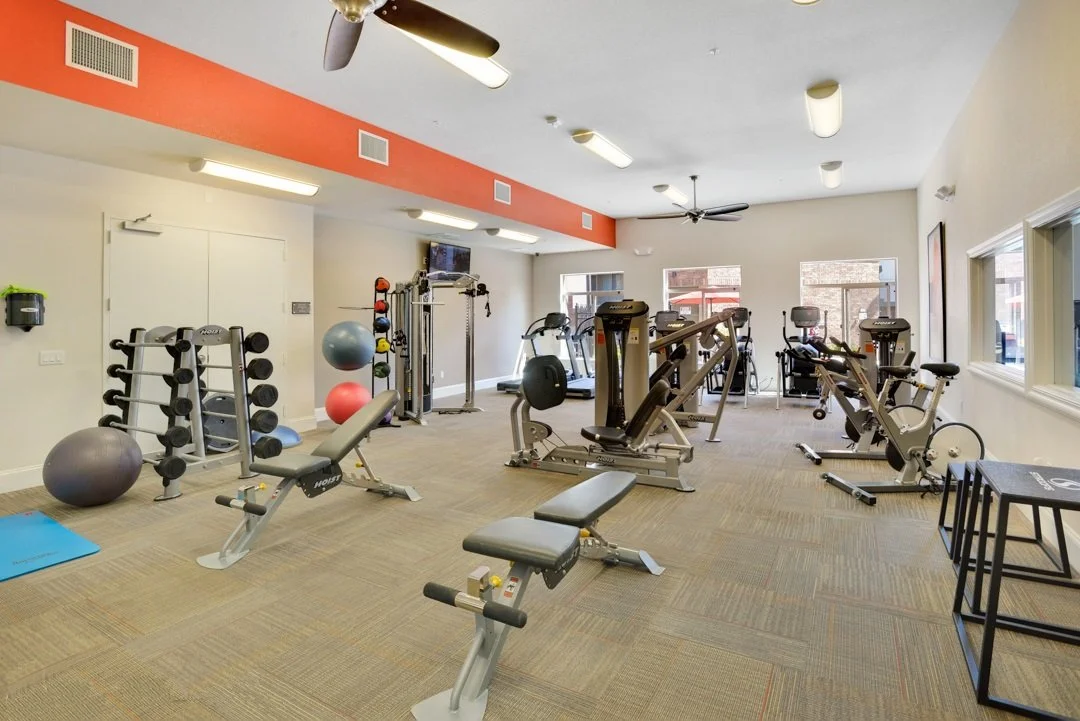 A well-equipped fitness room with various exercise machines, free weights, exercise balls, and mats. Large windows let in natural light, and ceiling fans are mounted on the ceiling.