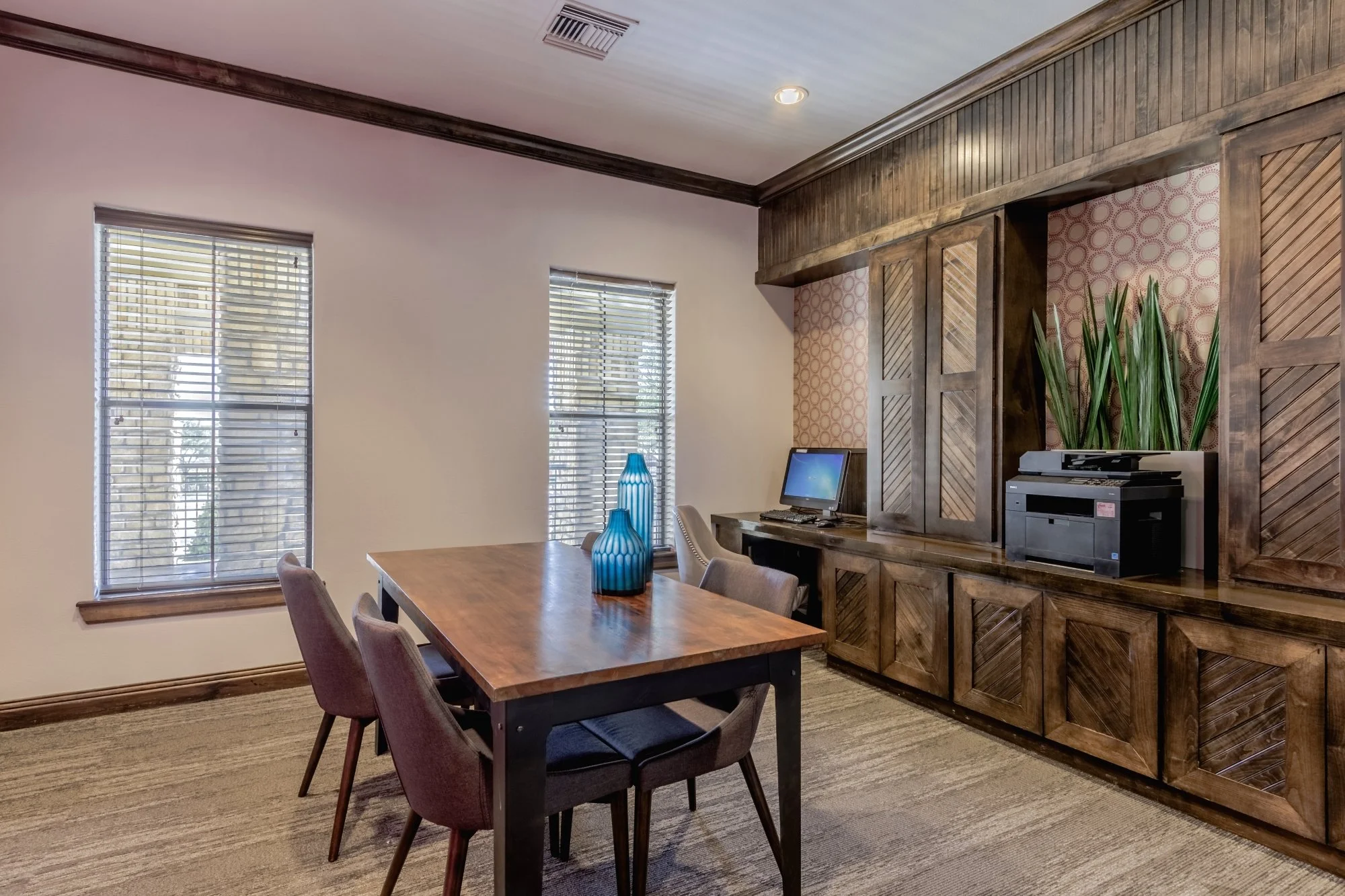 A dining room with a wooden table, four chairs, and two blue vases; a built-in wooden cabinet with plants and a printer; two windows with blinds; carpeted floor; beige and brown color scheme.