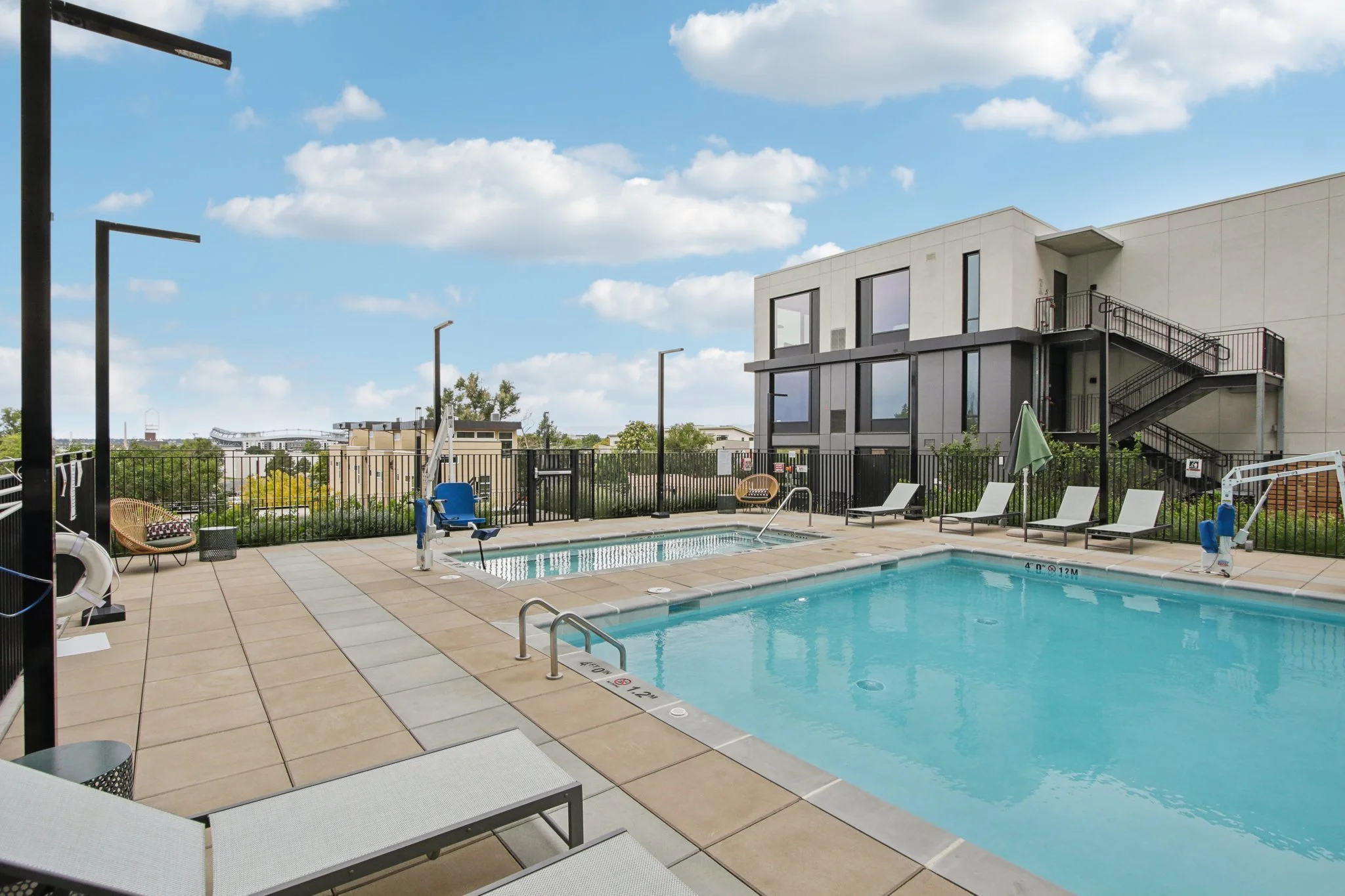 An outdoor swimming pool area with lounge chairs, a small hot tub, and modern apartment buildings in the background, under a partly cloudy sky.