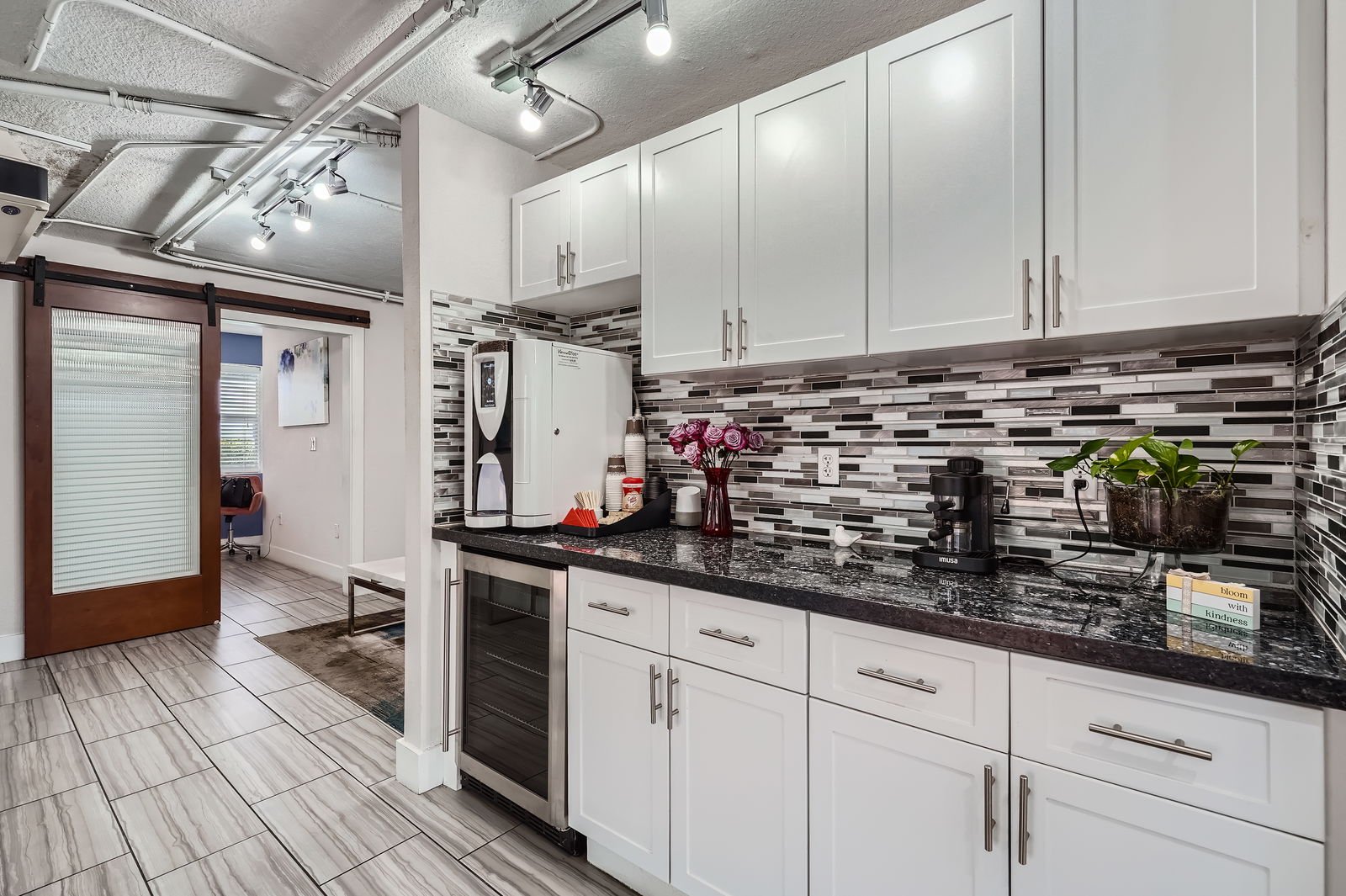 Kitchen with white cabinets, black granite countertop, mosaic tile backsplash, coffee maker, potted plant, flowers, and a wine refrigerator.