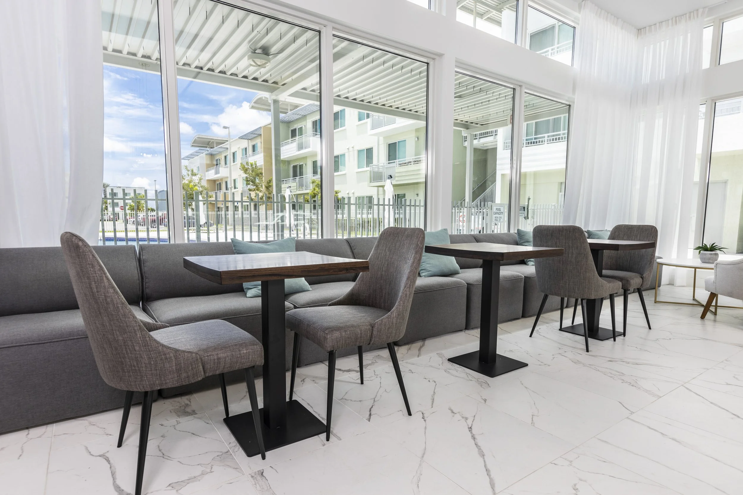 Interior of a modern lounge area with large windows, gray sofa, four chairs, wooden tables, and decorative pillows, overlooking an apartment complex with a pool outside.