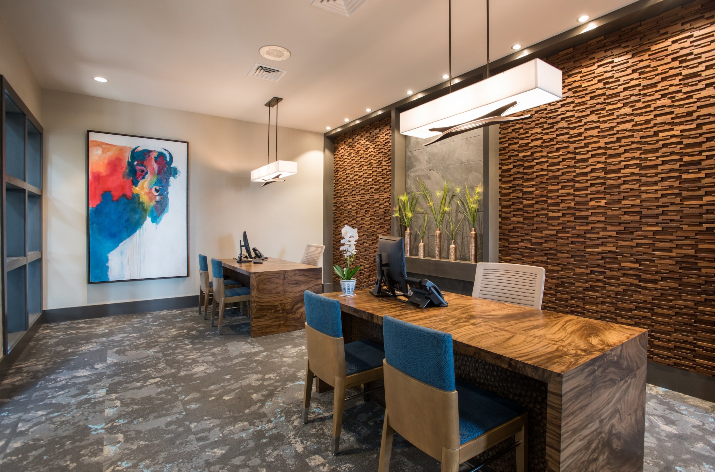 Modern hotel reception area with a large colorful dog painting on the wall, wooden desks, blue-upholstered chairs, and decorative plants with tall vases and greenery.