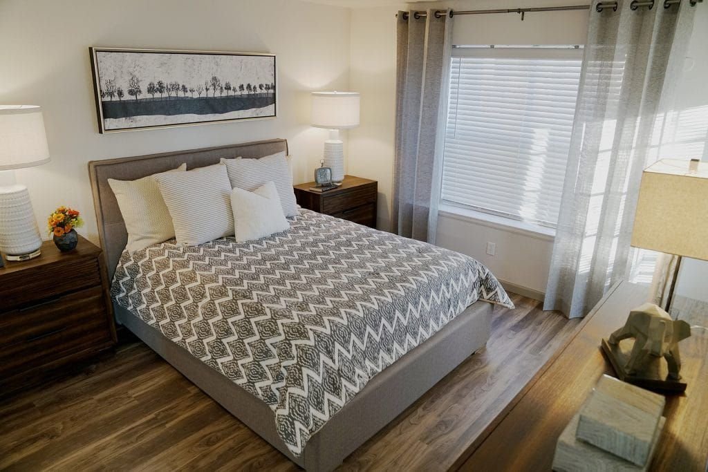 A cozy bedroom with a large bed featuring a patterned gray and white quilt, four pillows, wooden nightstands with lamps on each side, a framed landscape painting above the bed, a large window with sheer curtains, and a wooden dresser with decorative 