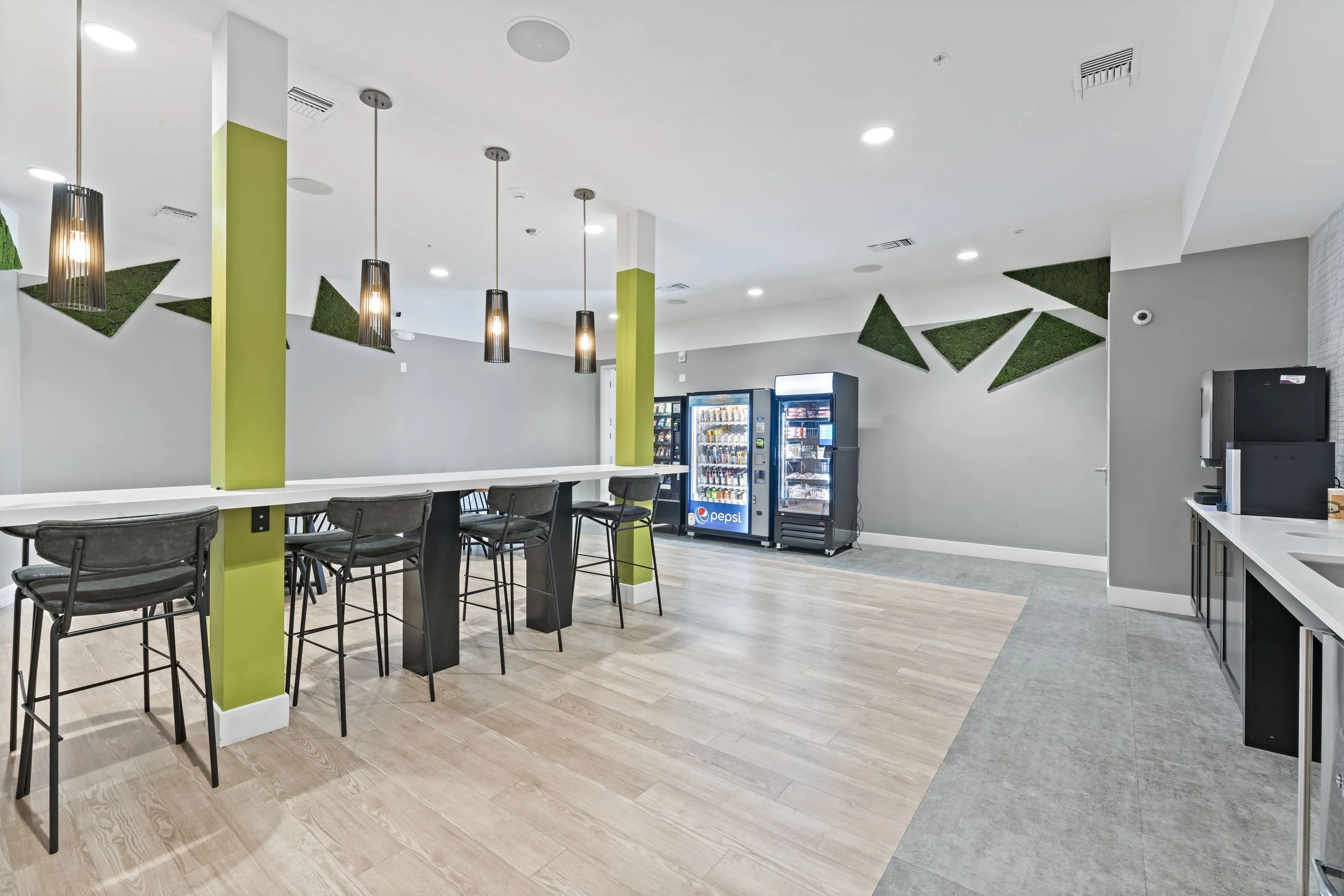 Empty lounge area with high table and chairs, vending machines, and a coffee station with a sink on the right, in a modern room with gray walls and decorative green wall accents.
