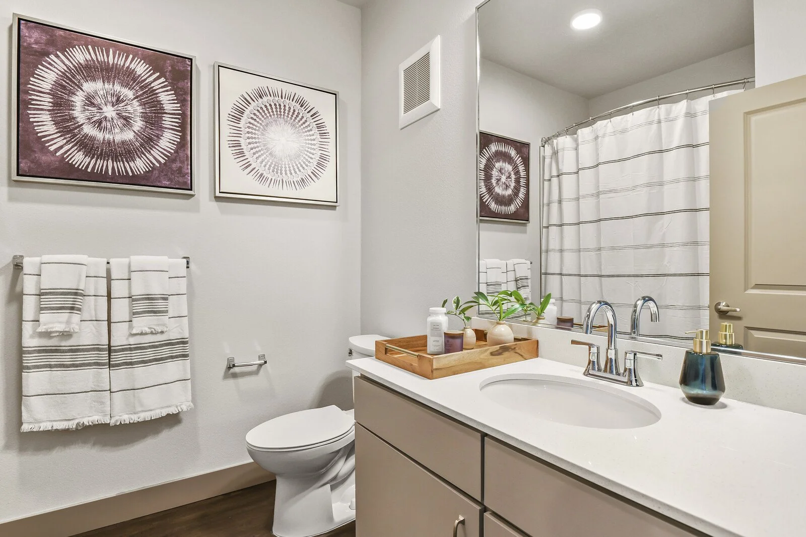 A modern bathroom with white walls, framed abstract art, a beige vanity with a white countertop, a plant in a small pot, and a soap dispenser. A mirror reflects the space, showing a shower with a striped curtain and a toilet with a tray of toiletries