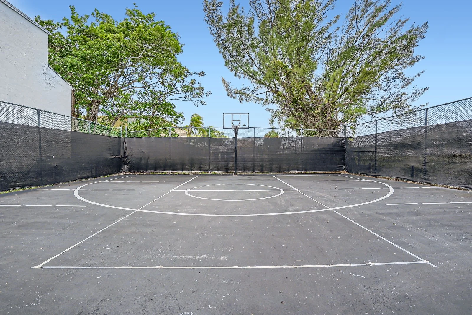 Empty outdoor basketball court with black fencing on all sides, large trees in the background, and a clear blue sky.