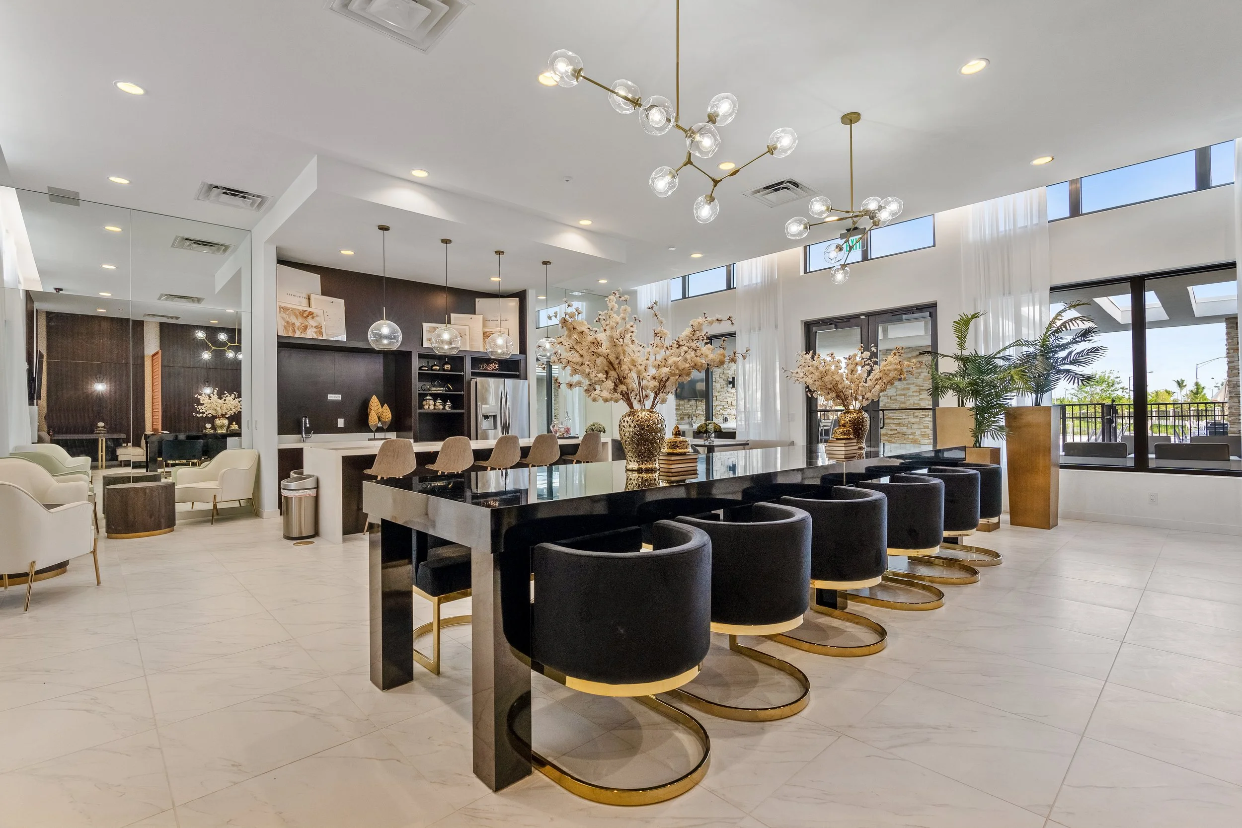 Modern, open-concept living space with a large black dining table, black velvet chairs with gold bases, a kitchen area with beige bar stools, and bright natural light coming through large windows, decorated with floral centerpieces and indoor plants.