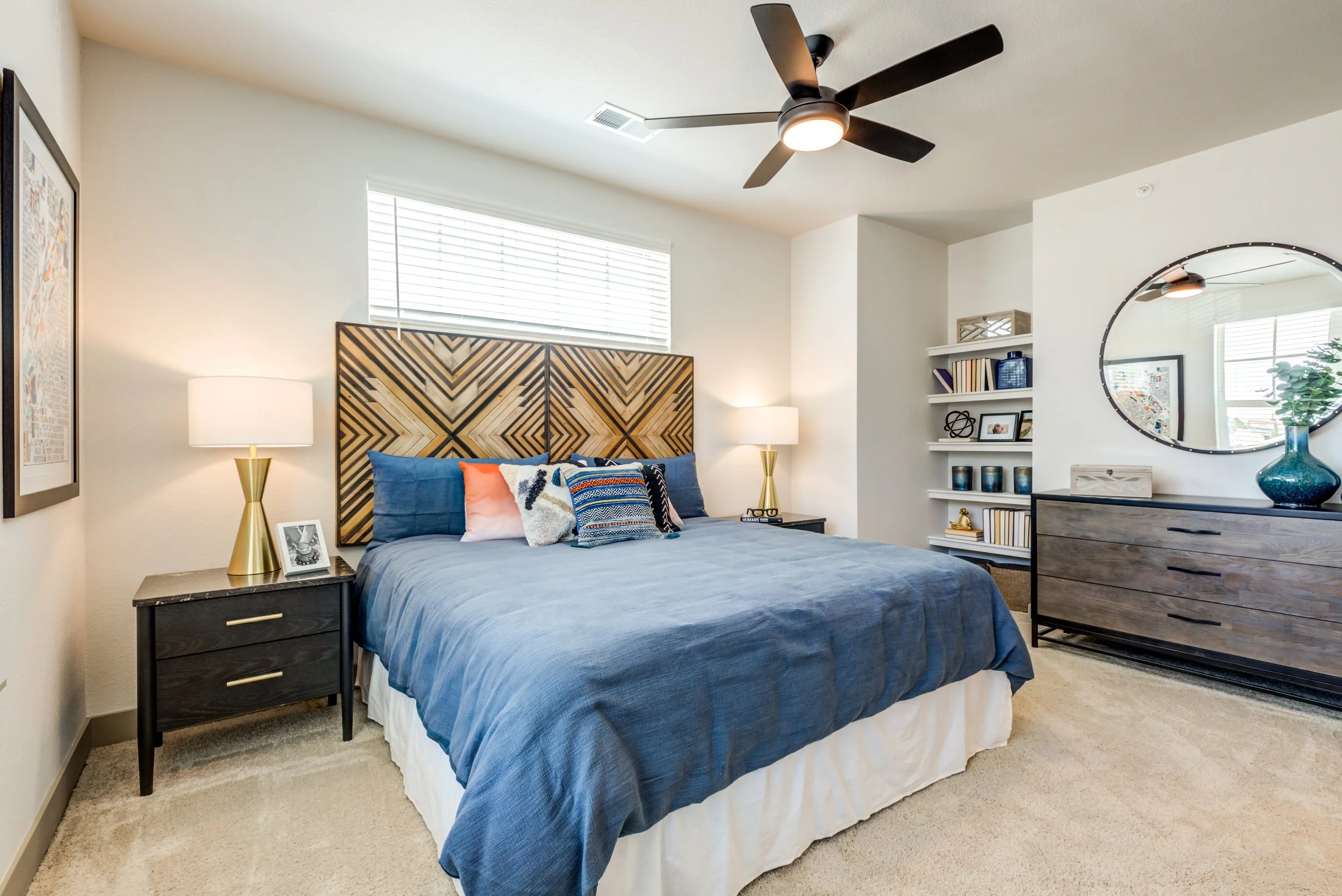 A modern bedroom with a large bed covered in a blue bedspread and decorative pillows. There are two black nightstands with gold lamps on each side of the bed, a ceiling fan above, a window with blinds, and a wooden dresser with a round mirror and dec