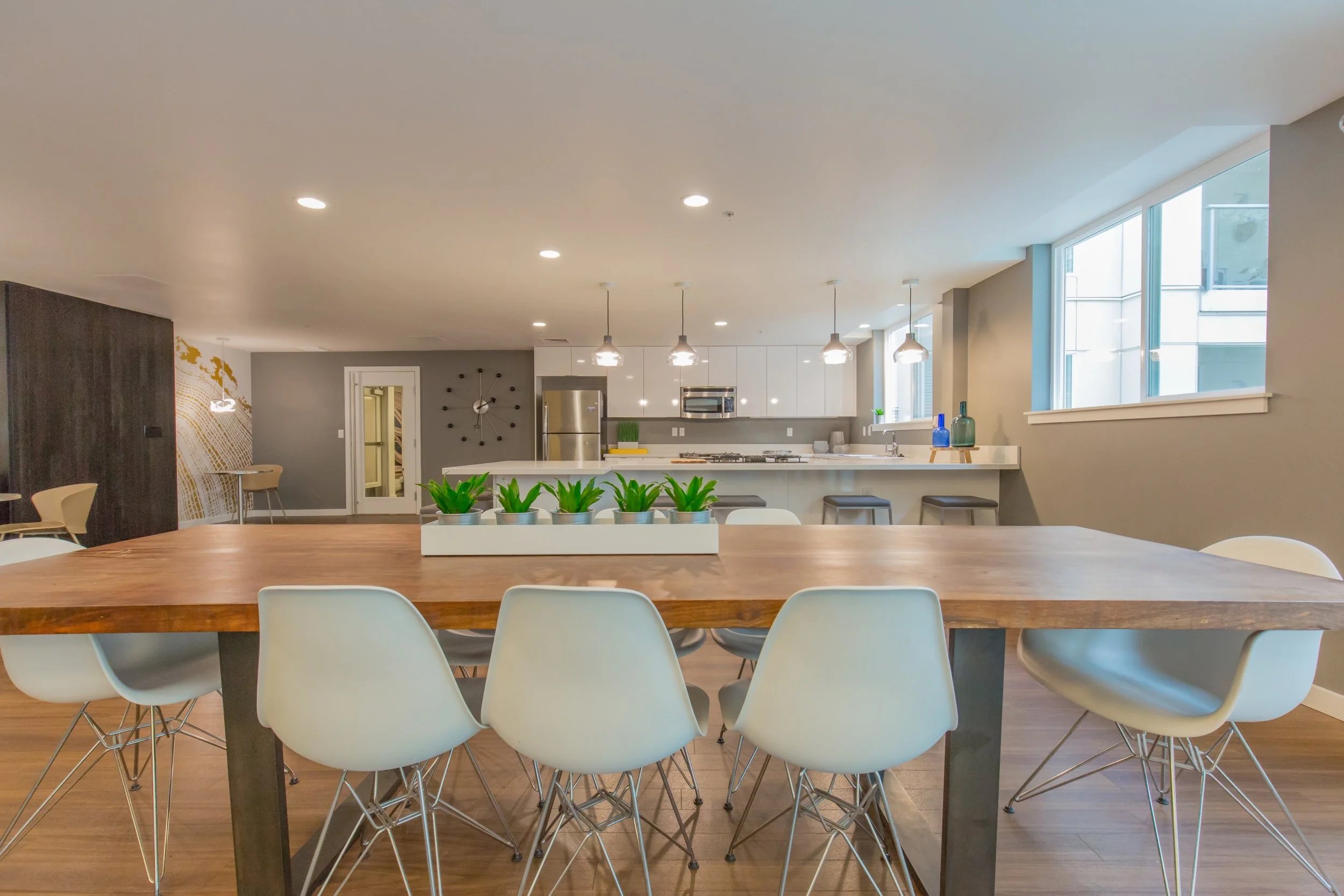 Modern open-concept kitchen and dining area with a large wooden table and white chairs, potted plants in the center, stainless steel appliances, pendant lighting, and large windows.