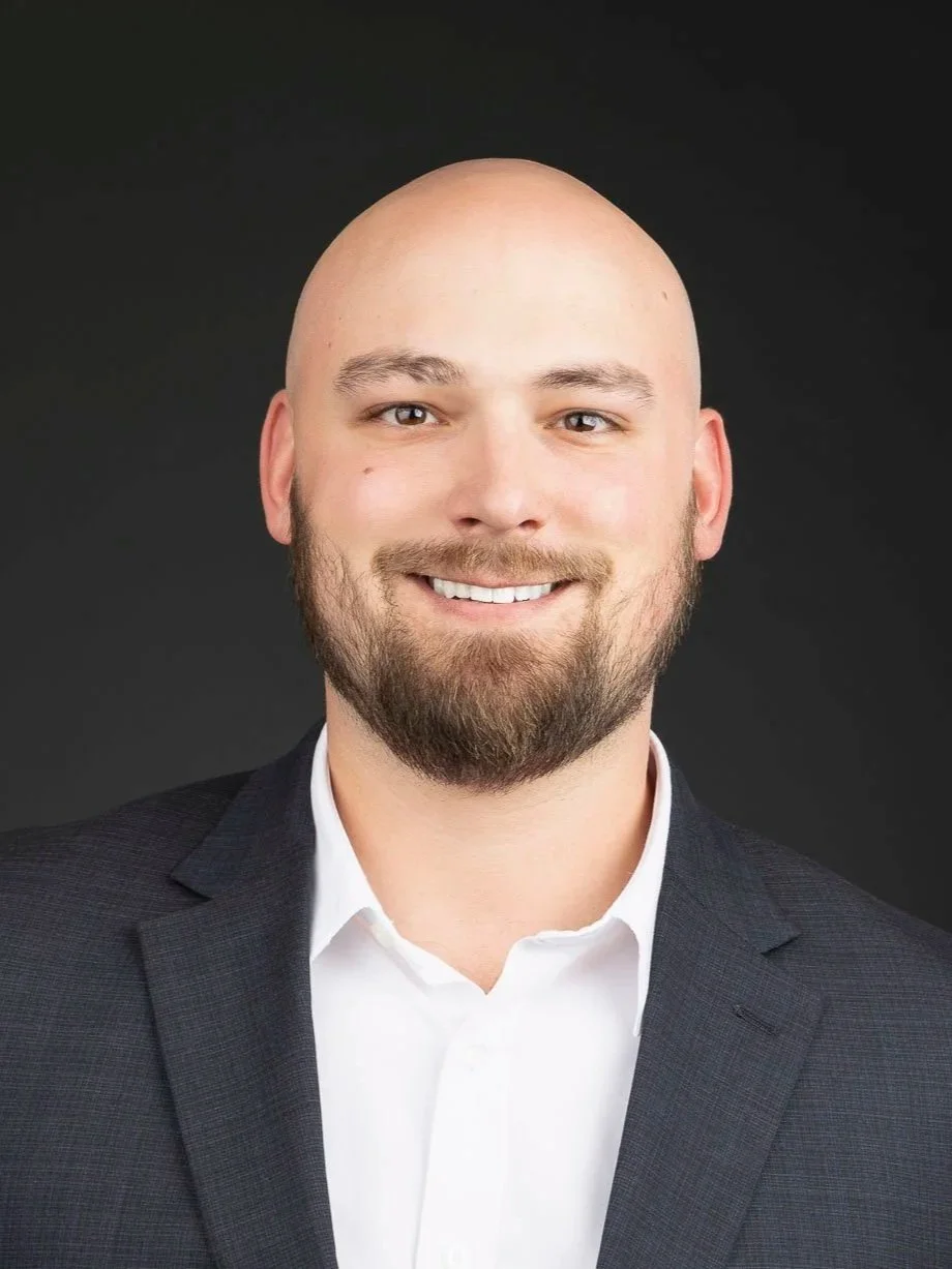 Headshot of a smiling man with a bald head, beard, wearing a dark suit jacket and white shirt against a black background.