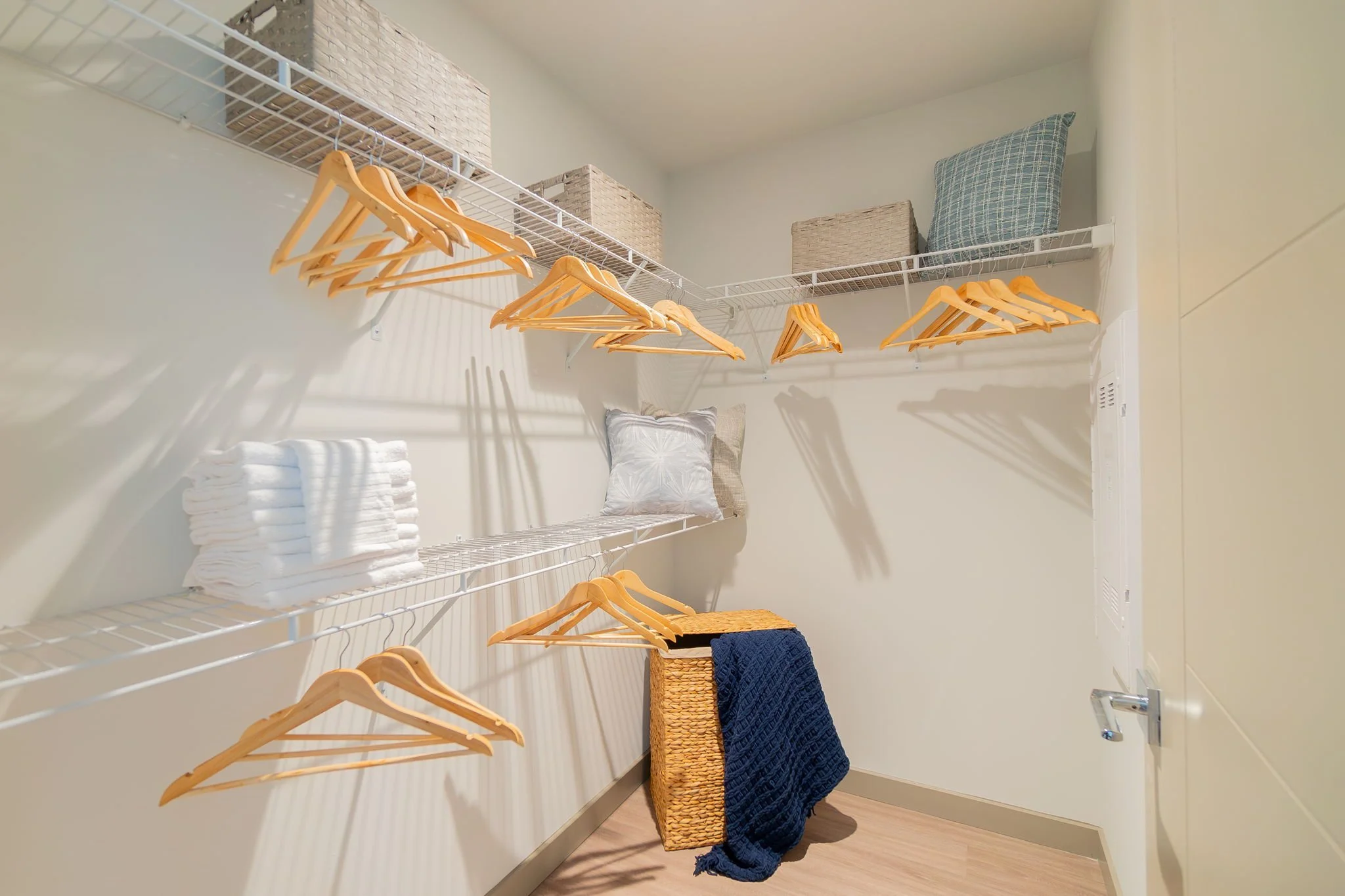 Empty walk-in closet with wire shelves, wooden hangers, and storage baskets, with folded towels and pillows, and a woven laundry basket with a blue blanket.