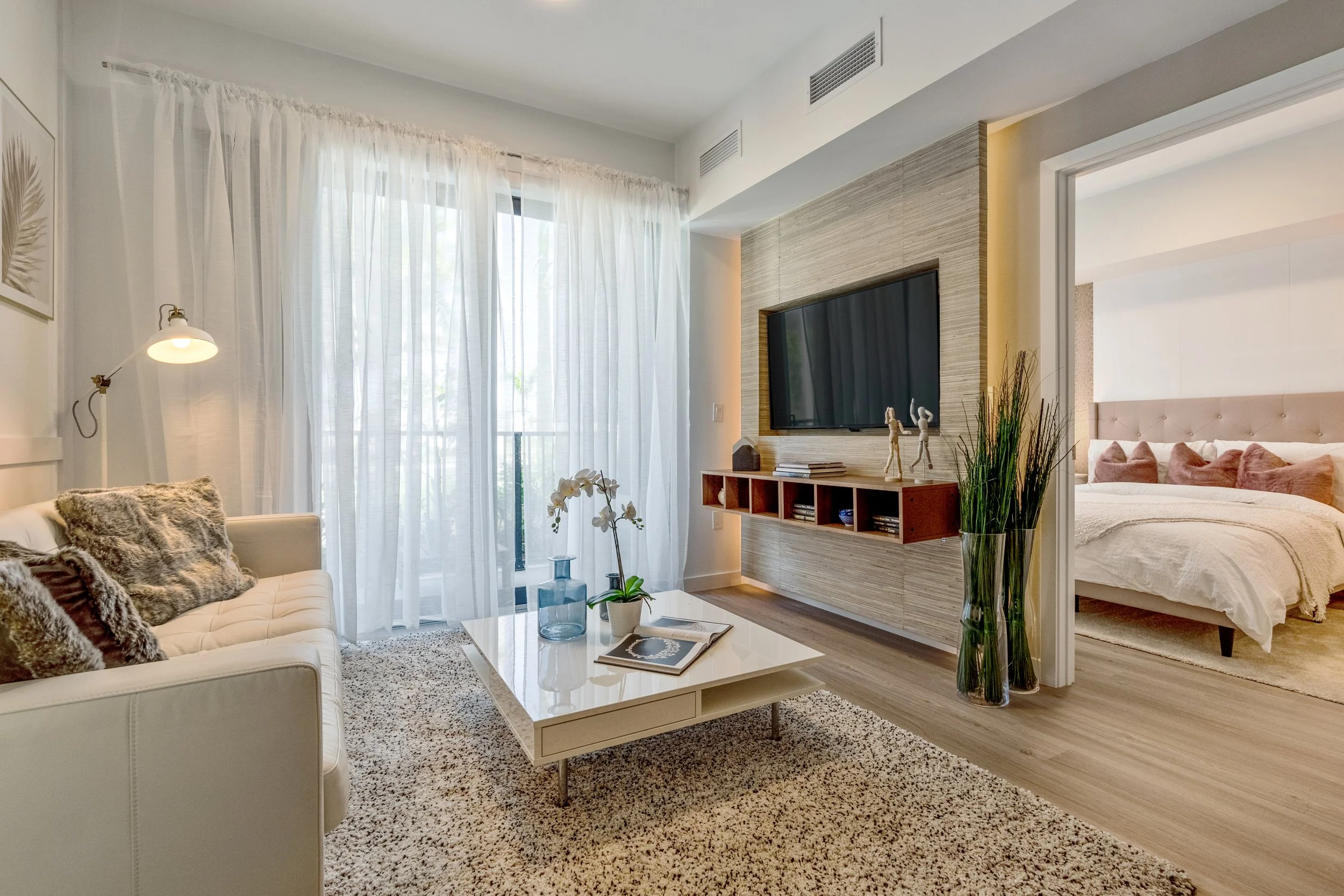 Living room with white sofa, fluffy pillows, a white coffee table, large window with sheer curtains, wall-mounted TV, wooden shelf with decorative items, a tall glass vase with green plants, and open doorway leading to a bedroom with a large bed and 