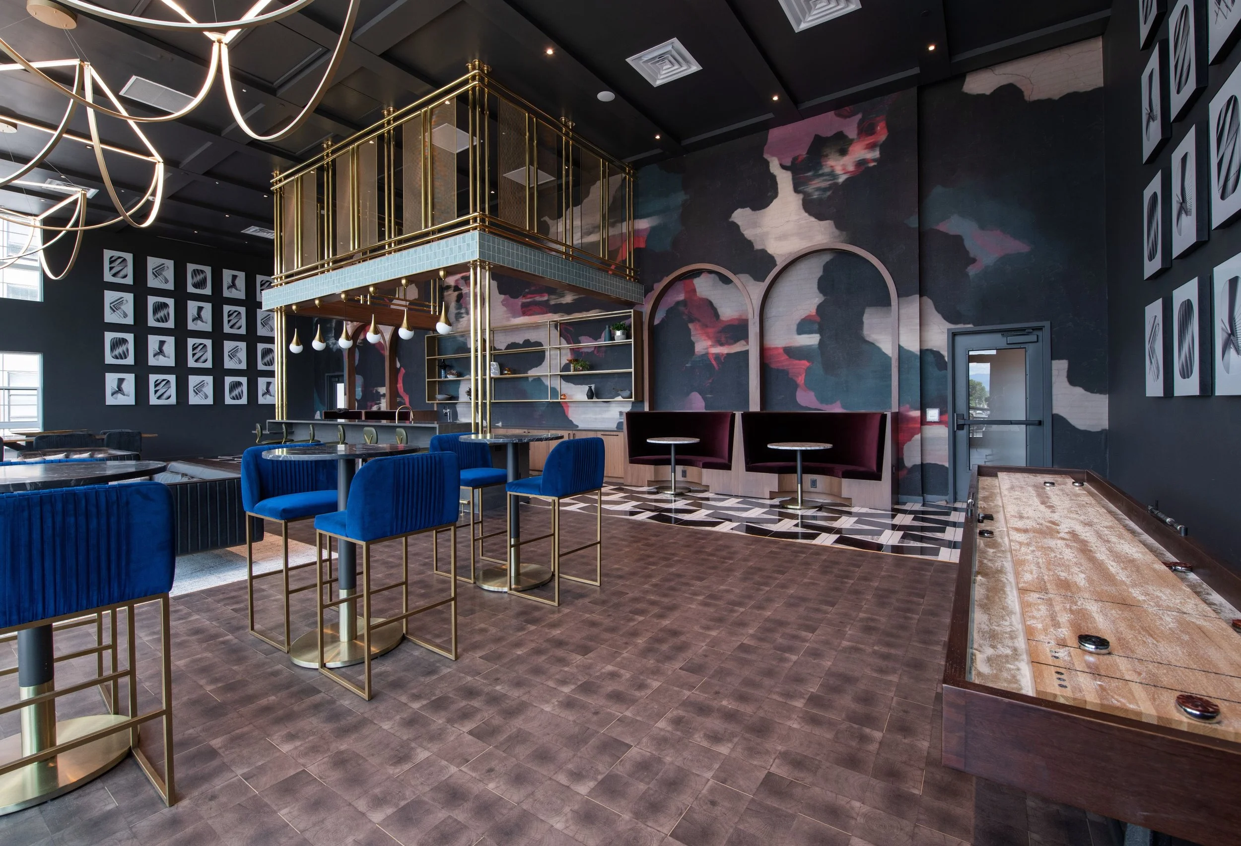 Interior of a modern entertainment or lounge area with dark walls featuring abstract art, blue velvet chairs, gold accents, a shuffleboard table, and billiard tables, illuminated by contemporary hanging light fixtures.