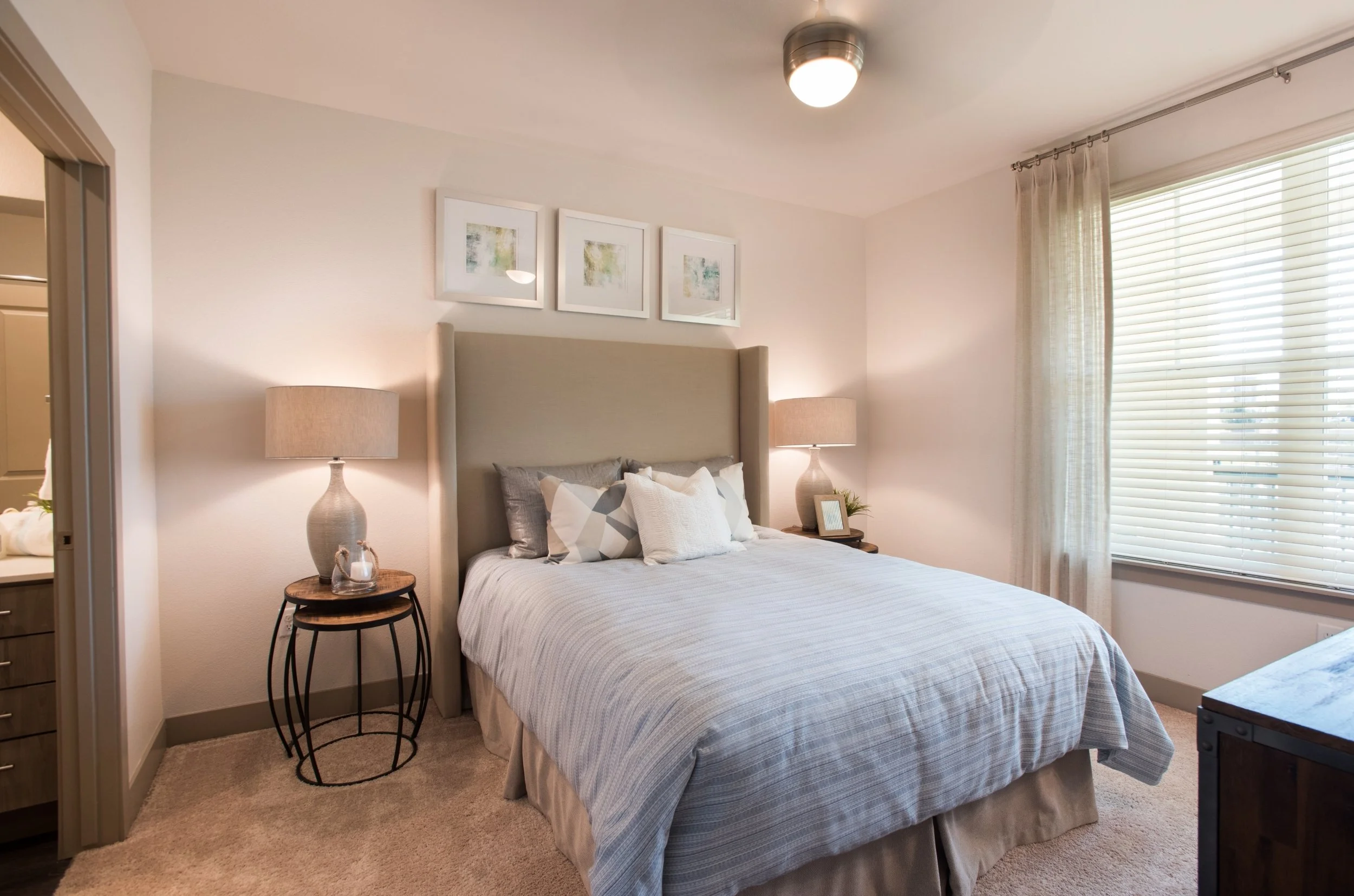 A cozy bedroom with a bed, beige headboard, and white bedding. Two nightstands with lamps are on each side of the bed, and a window with blinds and curtains is on the right. Artwork hangs above the headboard.