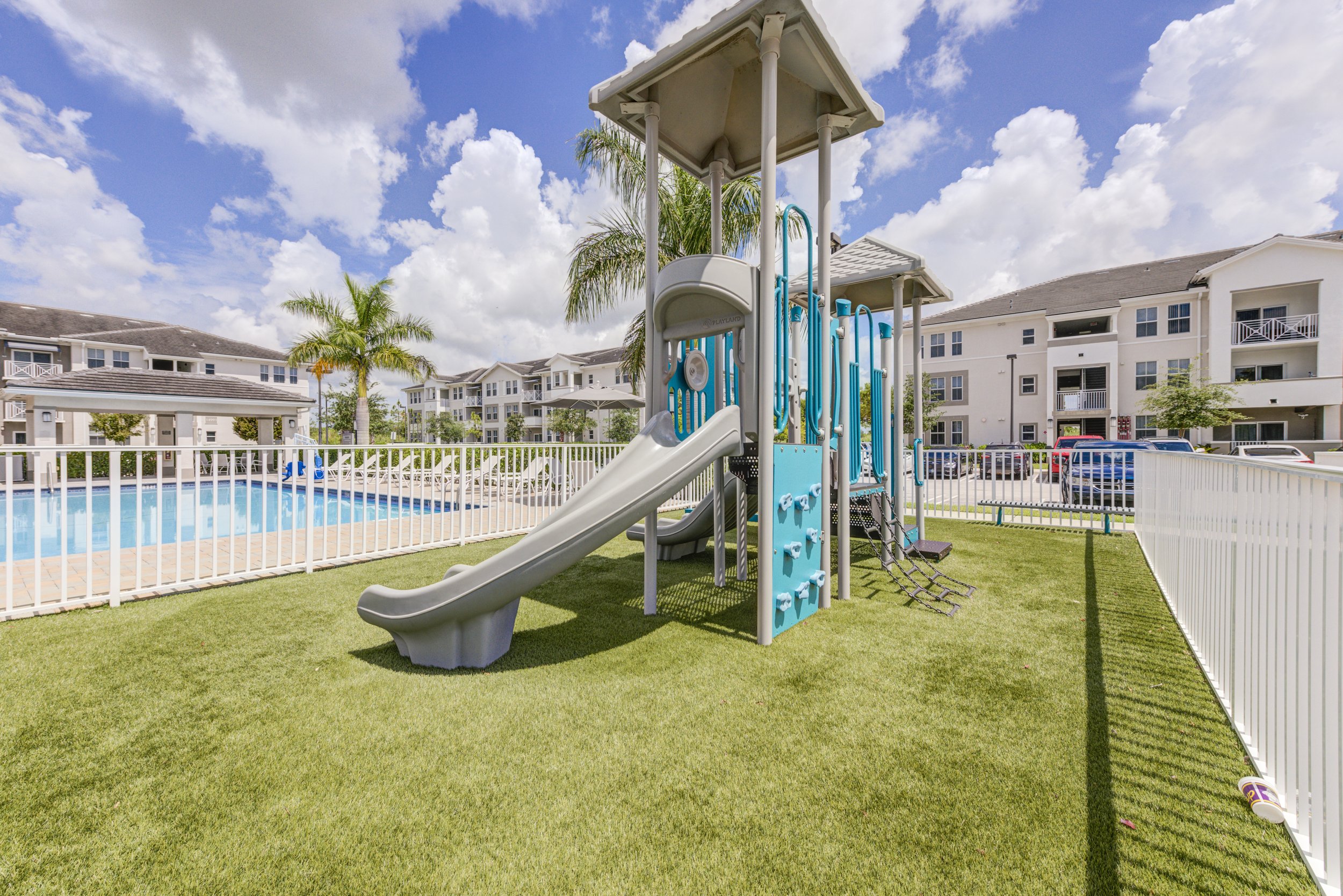 Child's playground with slide, climbing wall, and ladder, surrounded by a white fence, near a swimming pool with lounge chairs, in an apartment complex with multiple white buildings, palm trees, and a paved parking lot under partly cloudy blue sky.