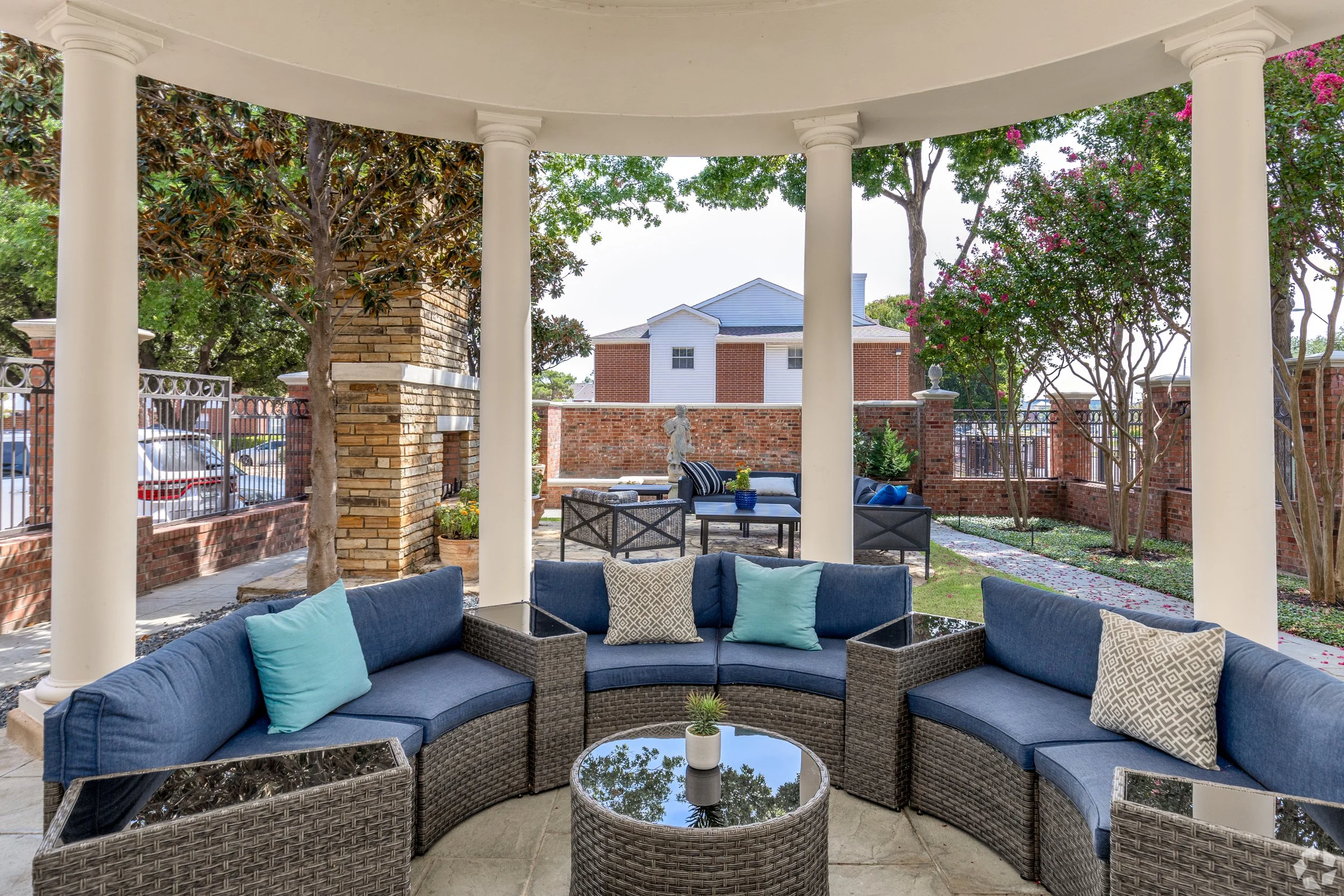 Outdoor patio with curved seating area with blue cushions and pillows, a round glass-topped coffee table, and surrounding trees and brick walls.