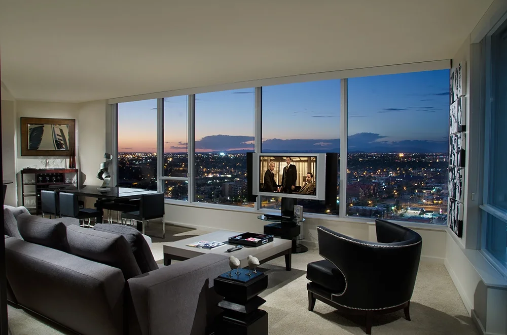 Living room with large window view of a cityscape at dusk, featuring a gray sofa, black armchair, and a TV showing a scene with three people.