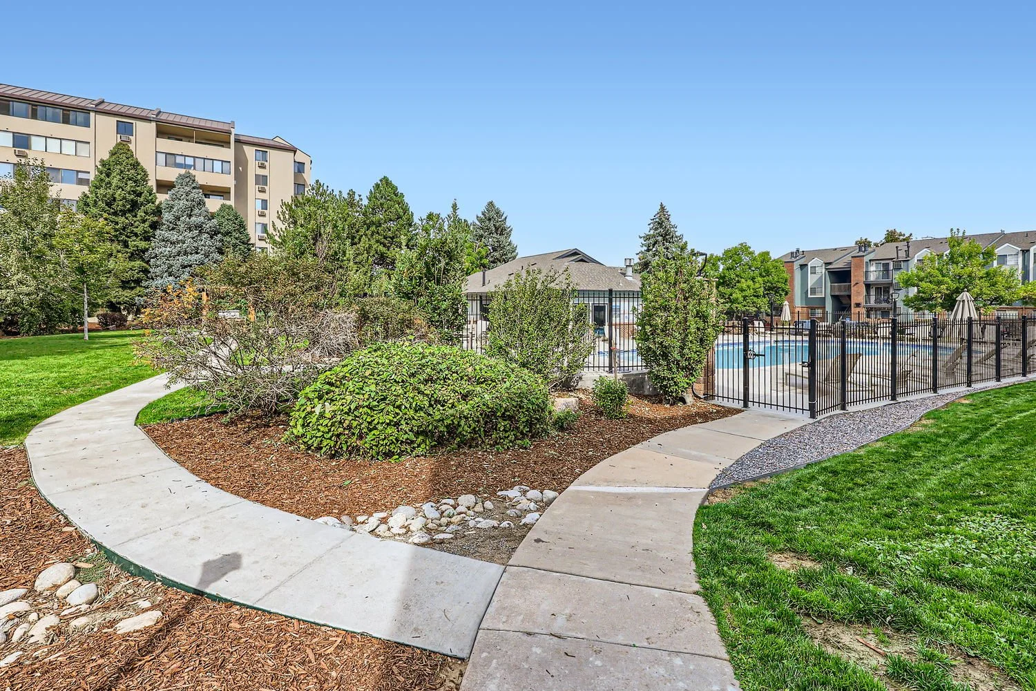 Pathways in a residential complex garden with grass, bushes, and trees, enclosed swimming pool with a black fence, surrounding apartment buildings, and a clear blue sky.