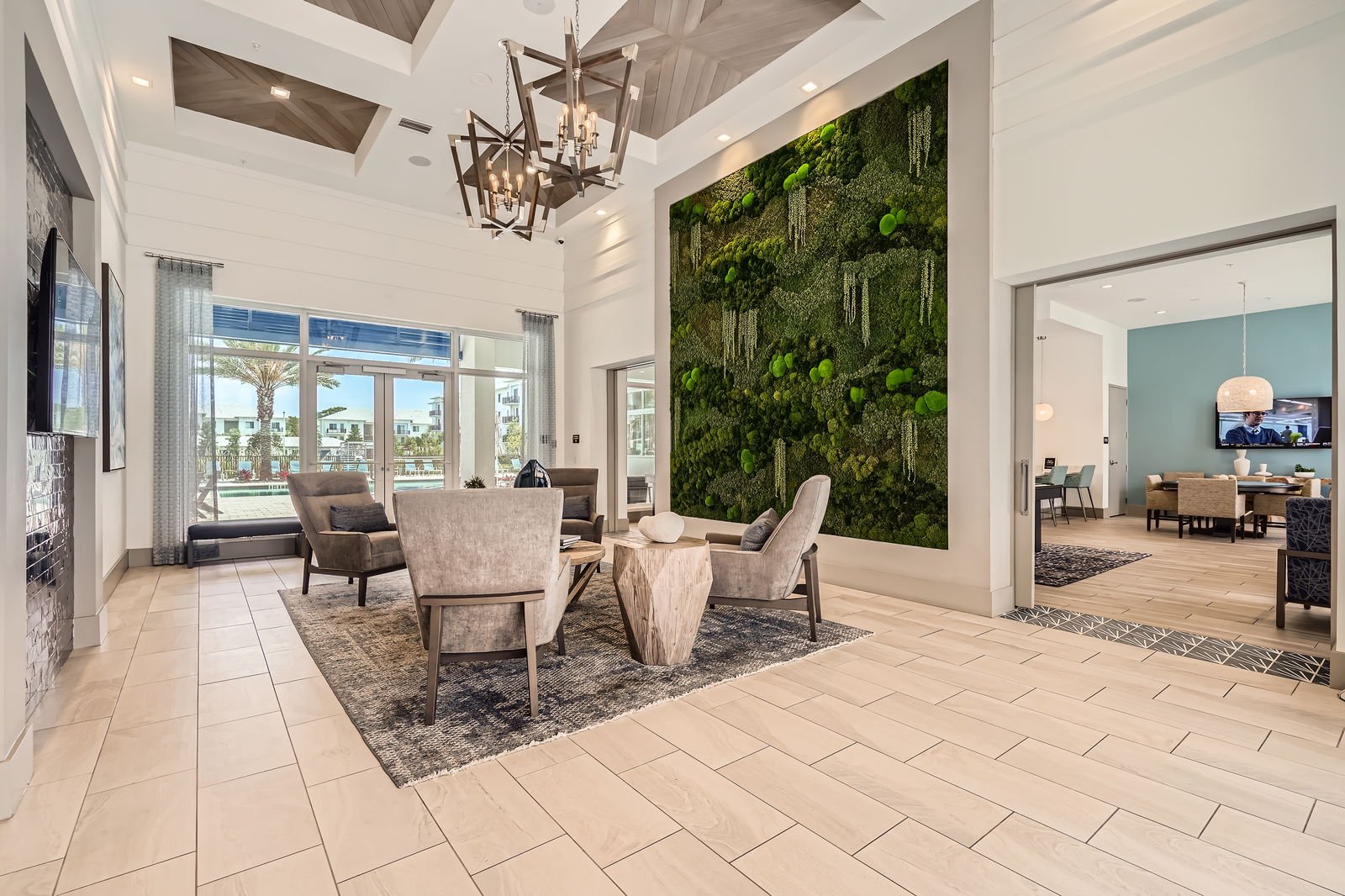 Modern living room with a large green wall art, beige chairs and a wooden table, large windows with curtains, tiled floor, and an adjacent room with a dining area.