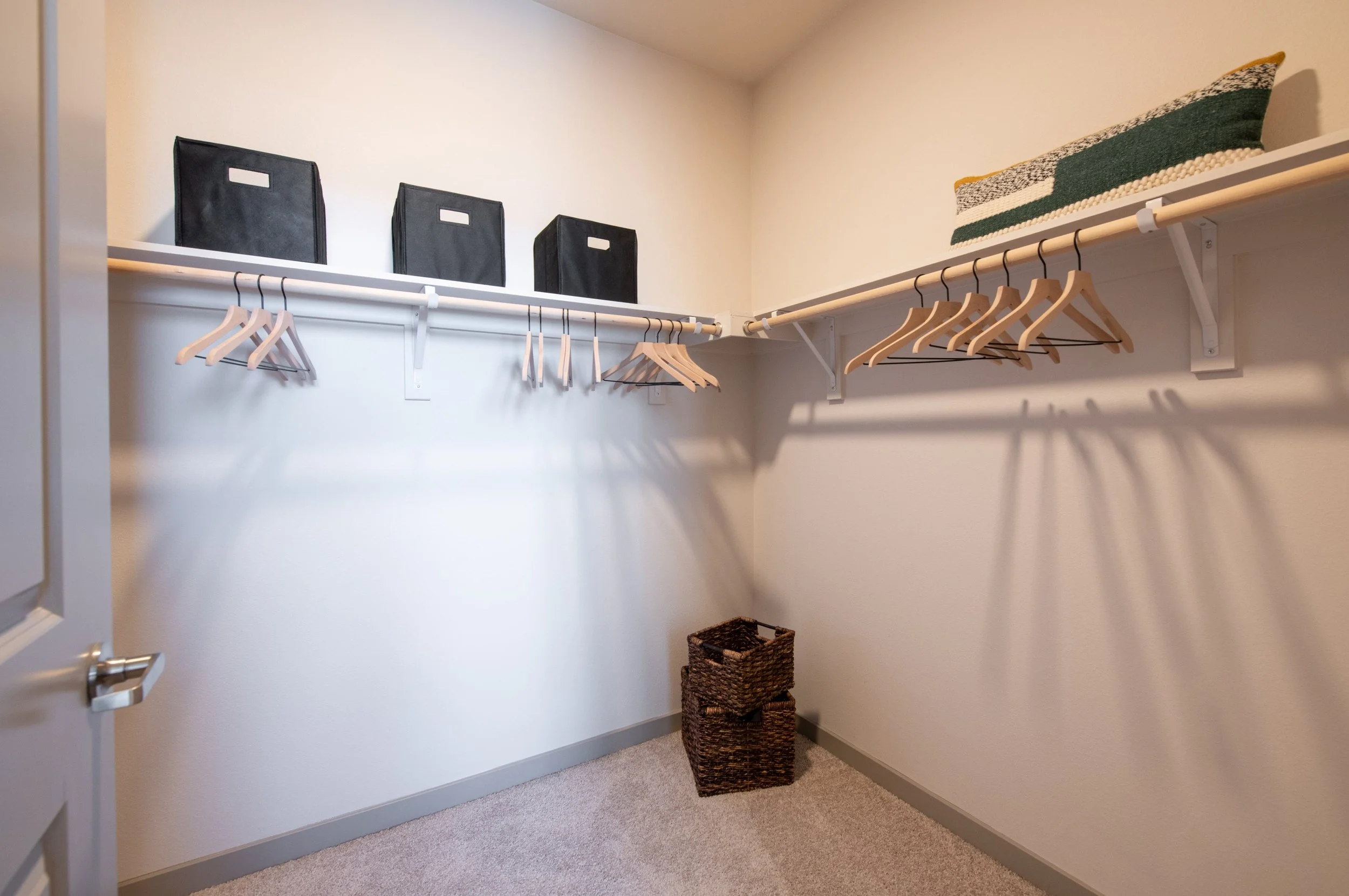 Empty walk-in closet with white shelves, black storage bins, wooden hangers, a woven basket, and a pillow on the top shelf.