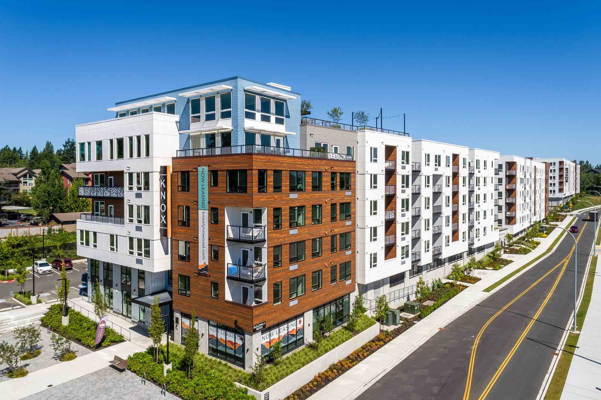 Modern multi-story apartment building with white, blue, and wood exterior finishes, large windows, and balconies, situated along a paved street with landscaped surroundings and parking areas.