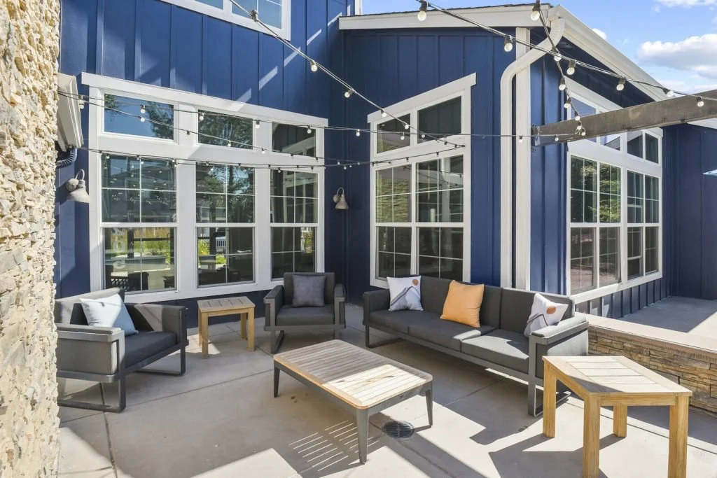 Outdoor patio area with gray seating, wooden tables, and string lights, in front of a blue house with large windows.