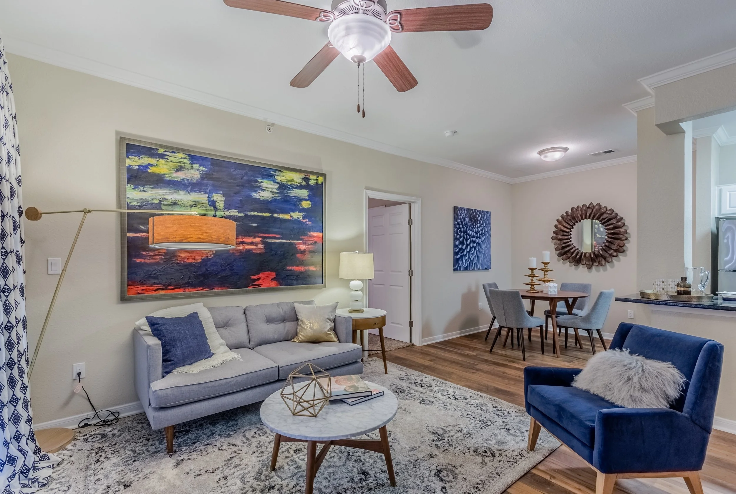 A living room with a beige wall, a light gray sofa with pillows, a round marble coffee table, a blue armchair with a fluffy pillow, a multi-colored pendant lamp, a wooden ceiling fan, a wood floor, a patterned carpet, art pieces on the wall, and a di