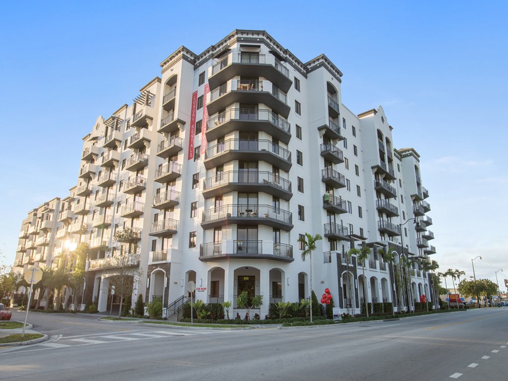A large white apartment building with multiple floors, balconies, and arched windows, situated at a street corner with palm trees and landscaping in the foreground.