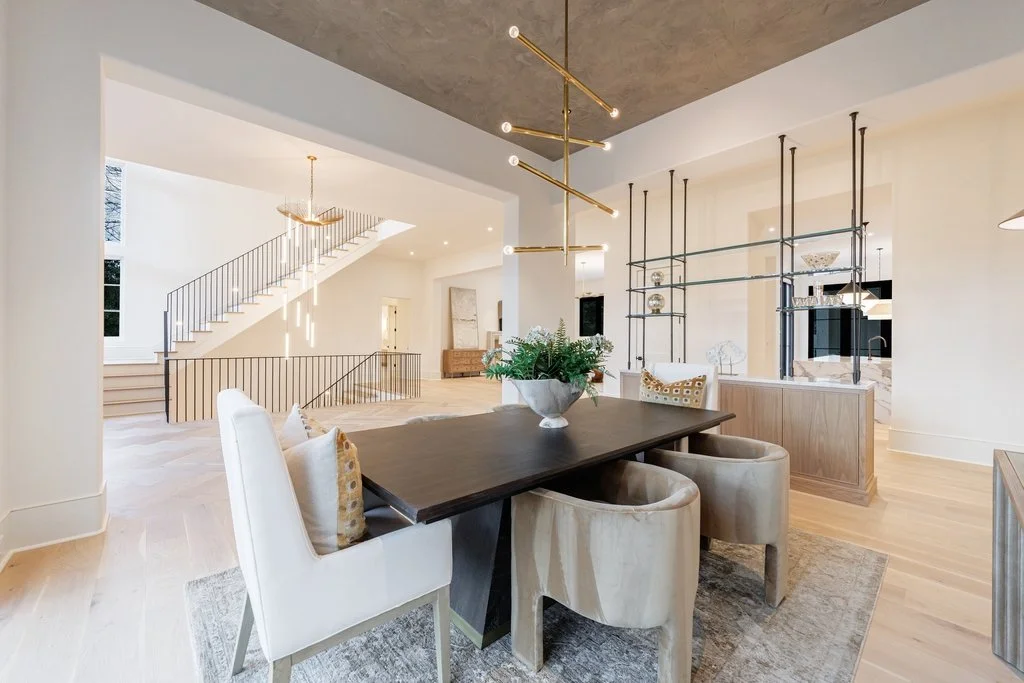 Open-concept dining and living area with light wood floors, modern chandelier, staircase with black railings, and neutral decor.