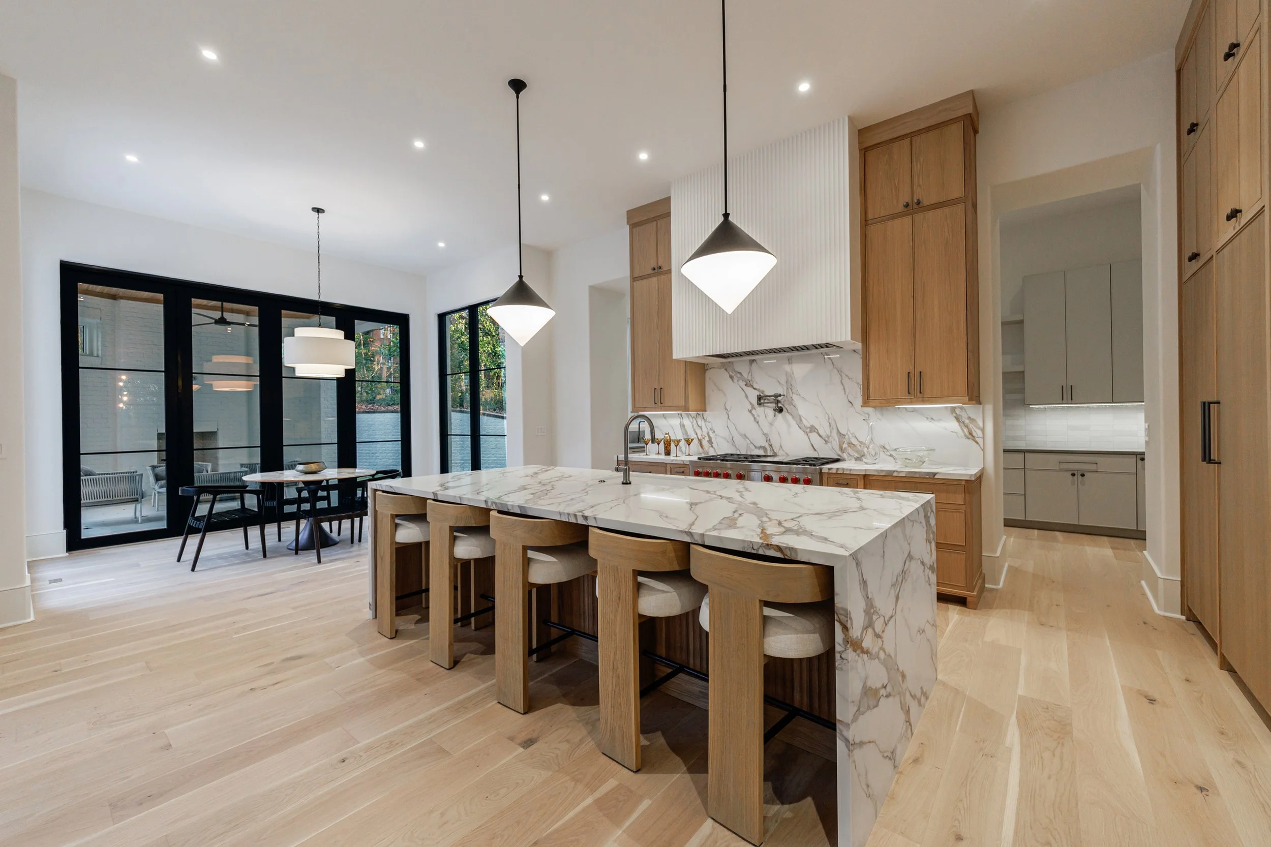 Modern kitchen with marble island, light wooden cabinets, and black pendant lights, adjacent to a dining area with large windows and black-framed glass doors.