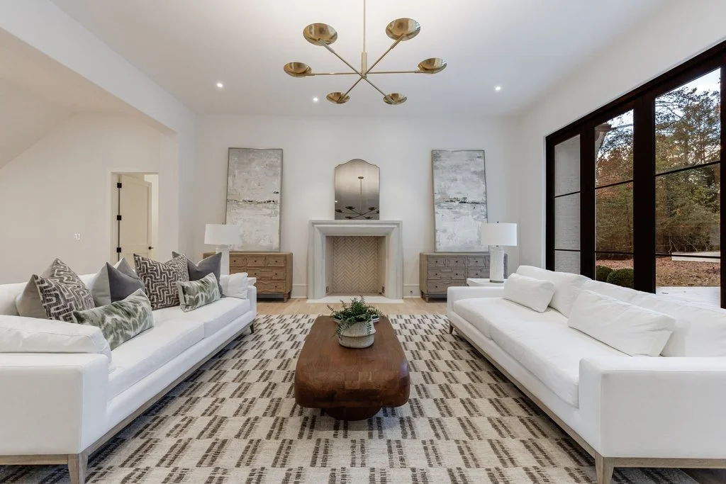 Modern living room with two white sofas, a wooden coffee table, abstract paintings, and large windows showing trees outside.