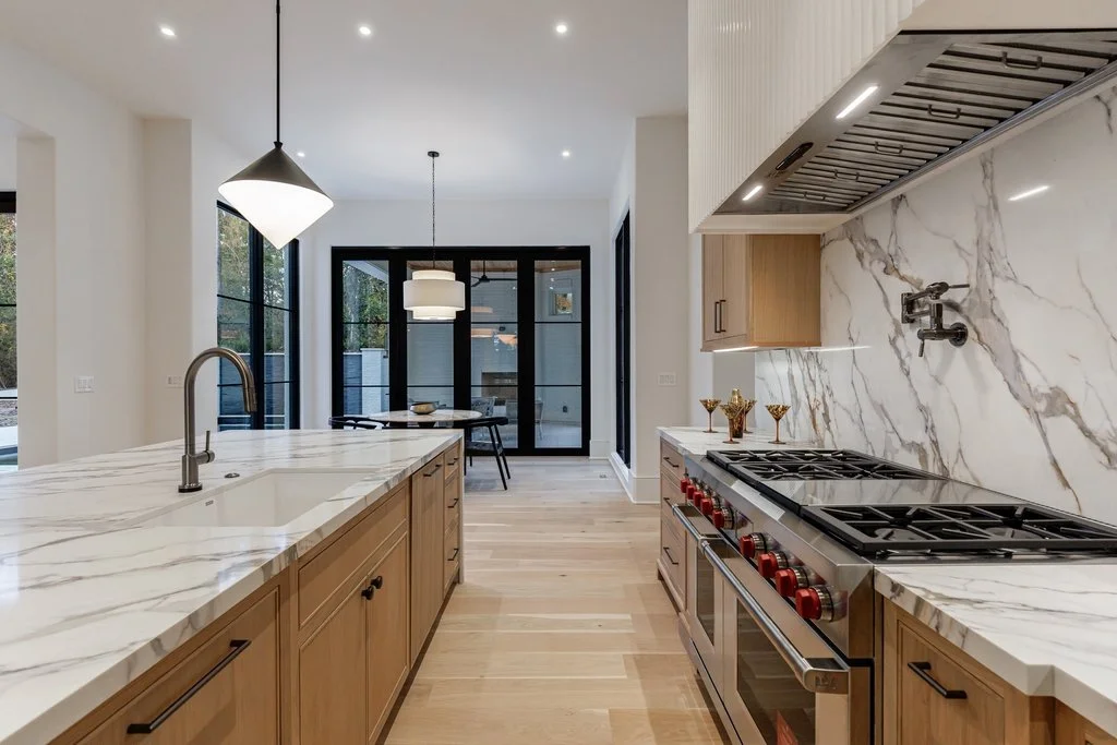 Modern kitchen with marble countertops, wooden cabinetry, stainless steel stove, and a dining area with hanging lights and large windows.