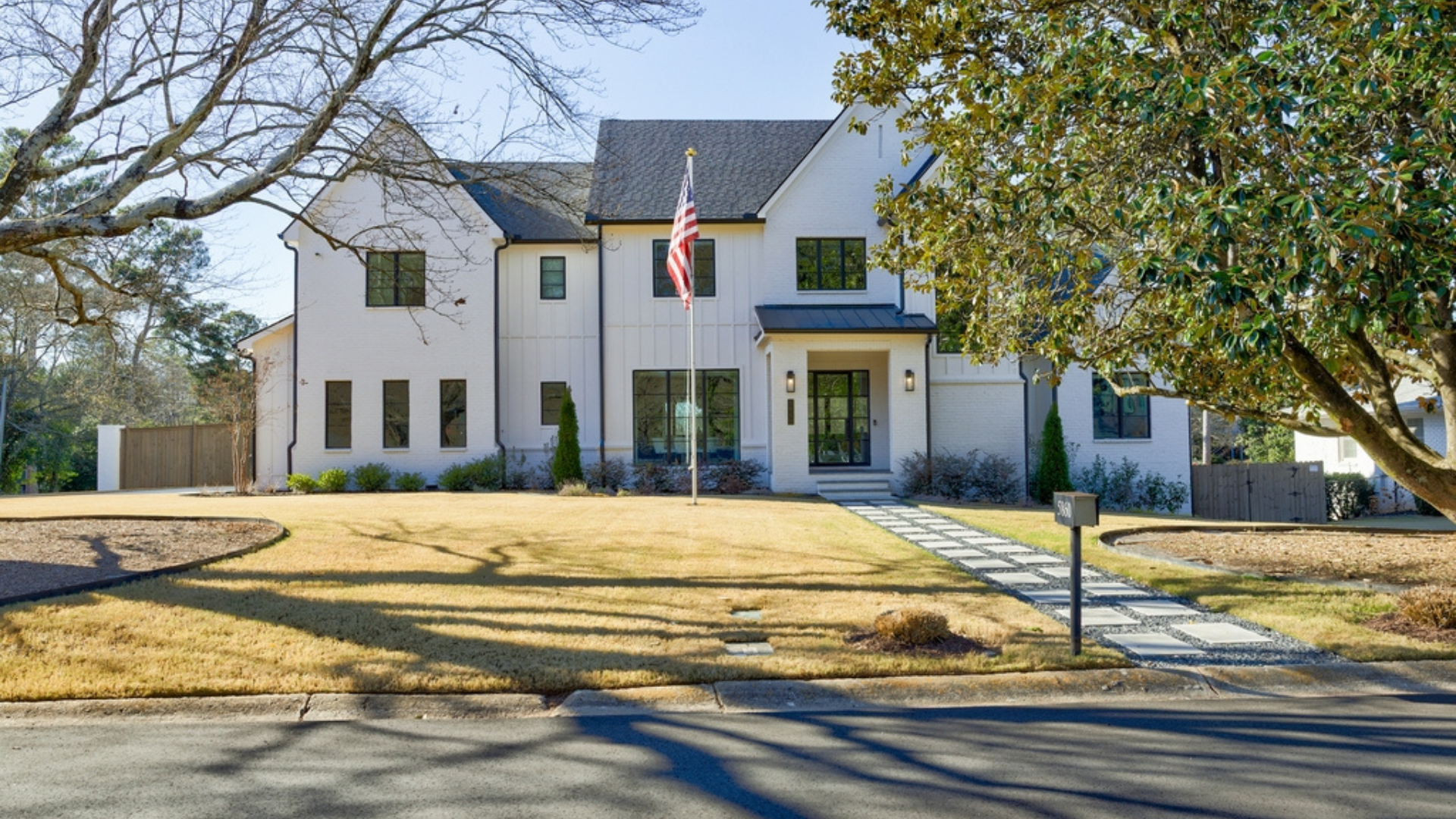 Front view of a modern white house with a dark roof, American flag, well-maintained lawn, stone walkway, and a large tree in the yard.
