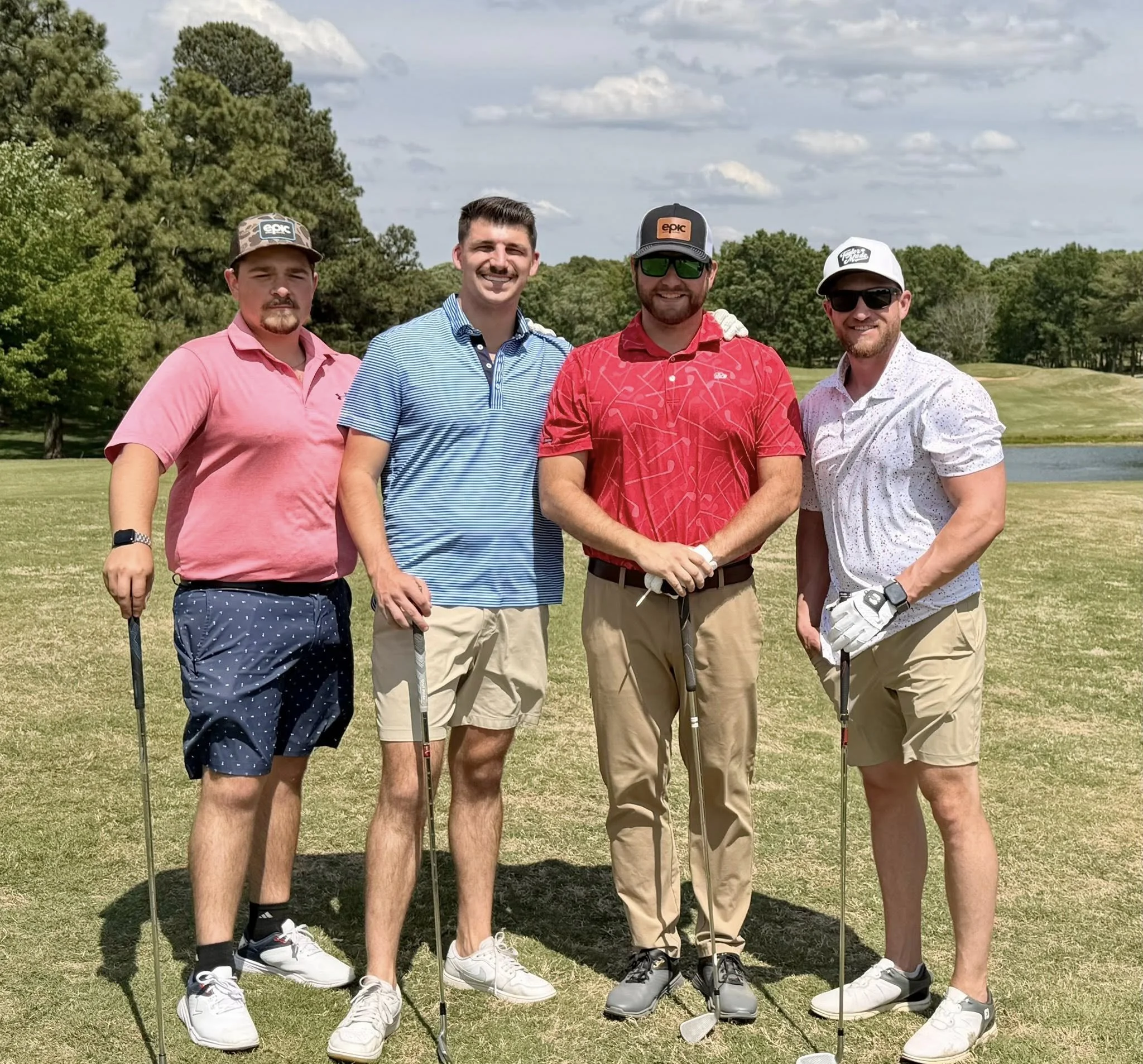 Group of four men standing on a golf course, holding golf clubs, dressed in casual golf attire, with trees and cloudy sky in the background.
