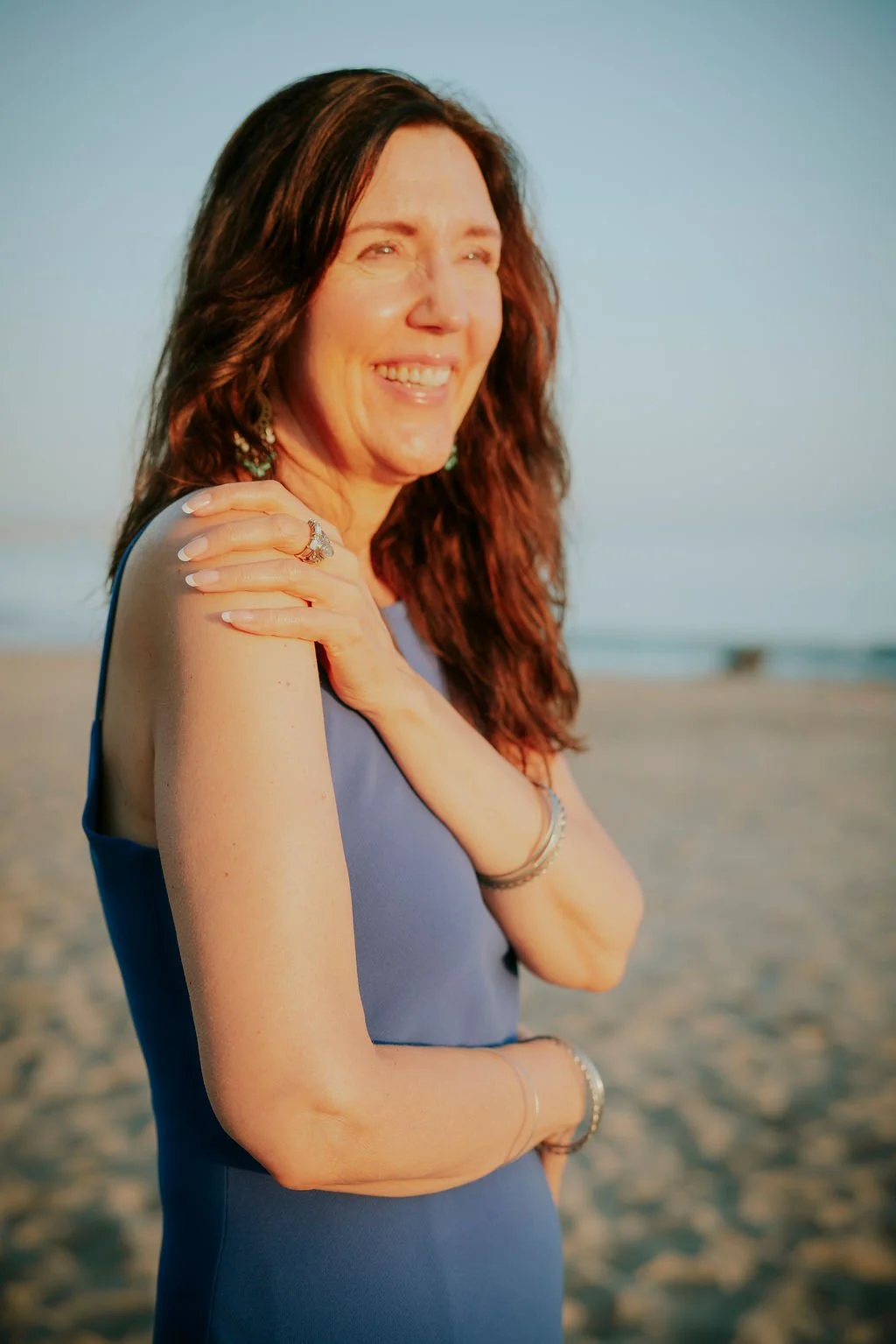 A woman smiling on a beach during sunset, with her hand on her shoulder and her eyes closed.