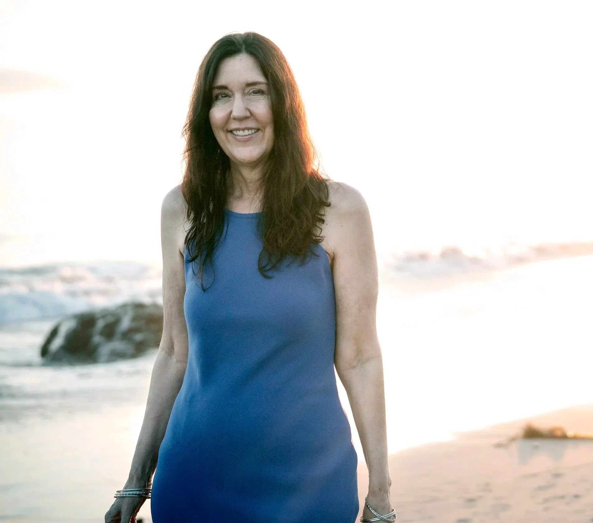 Woman with long dark hair smiling on a beach during sunset, wearing a blue sleeveless dress, with ocean waves in the background.