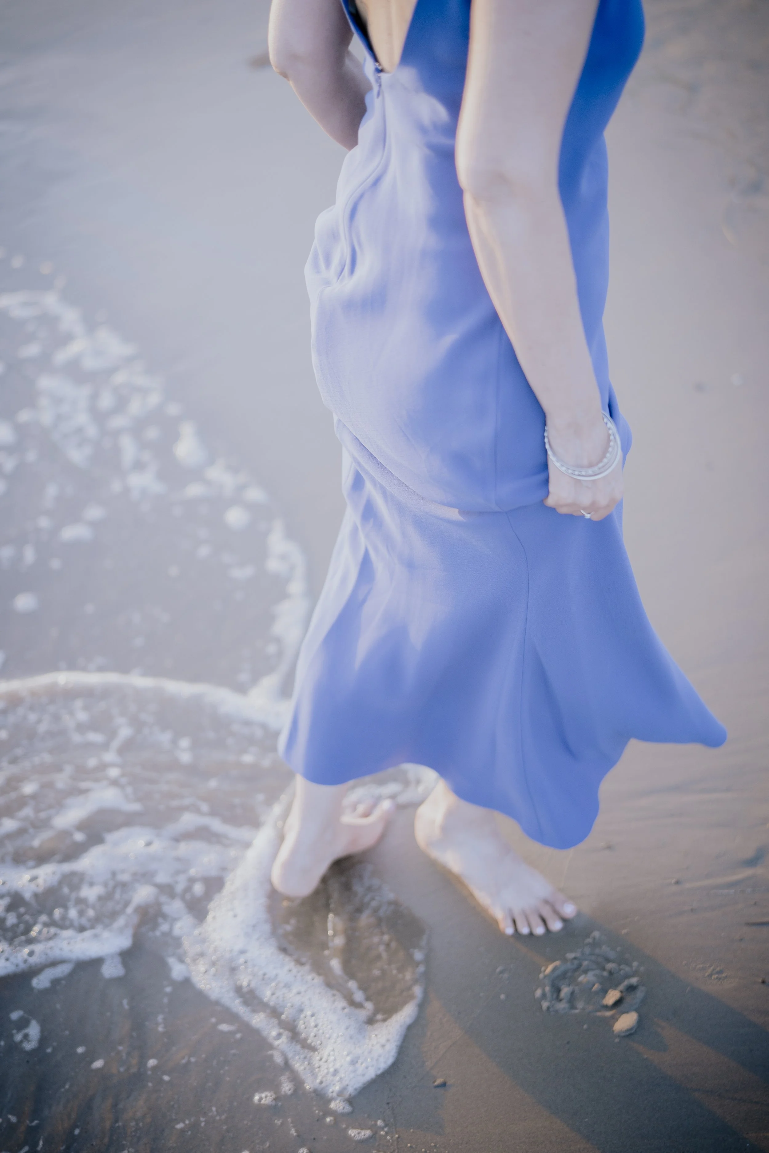 Person barefoot in a blue dress walking along the shoreline with waves touching their feet.