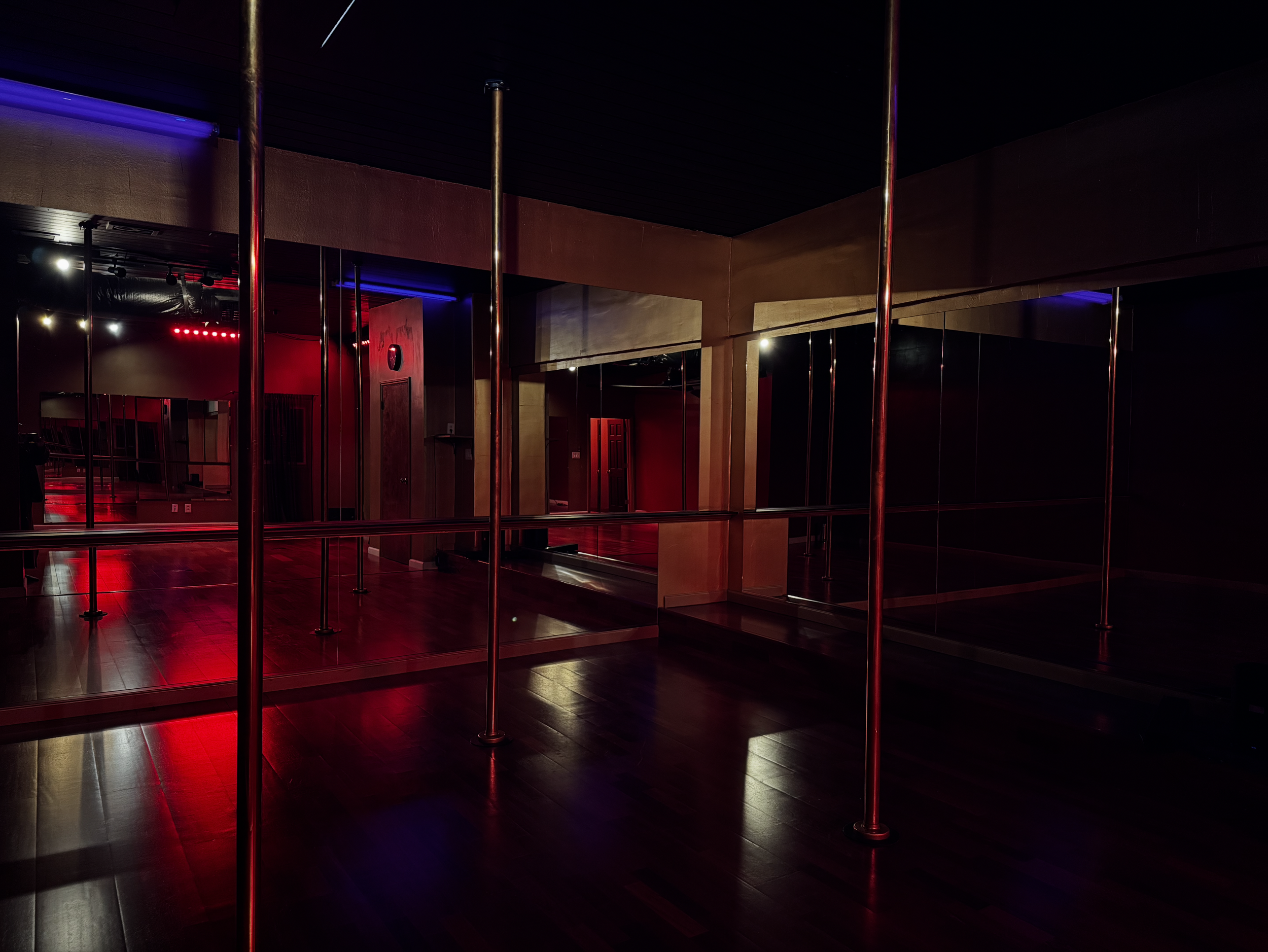 Dimly lit dance studio with mirrors, wooden floors, and metal ballet poles, illuminated by red and purple lighting.