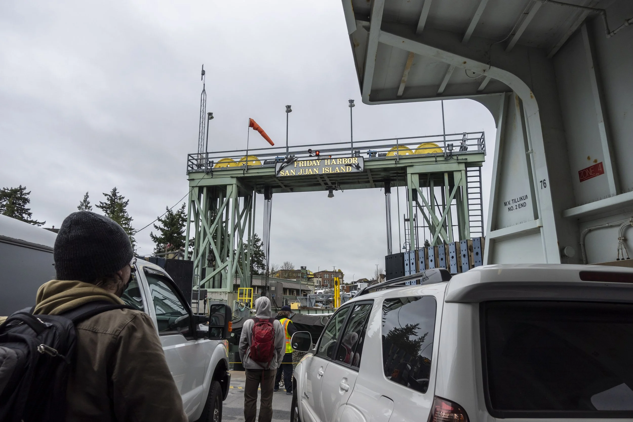 On ferry about disembark to Friday Harbor, WA. Looking from behind a couple of people and cars with the ferry terminal beyond on a grey sky