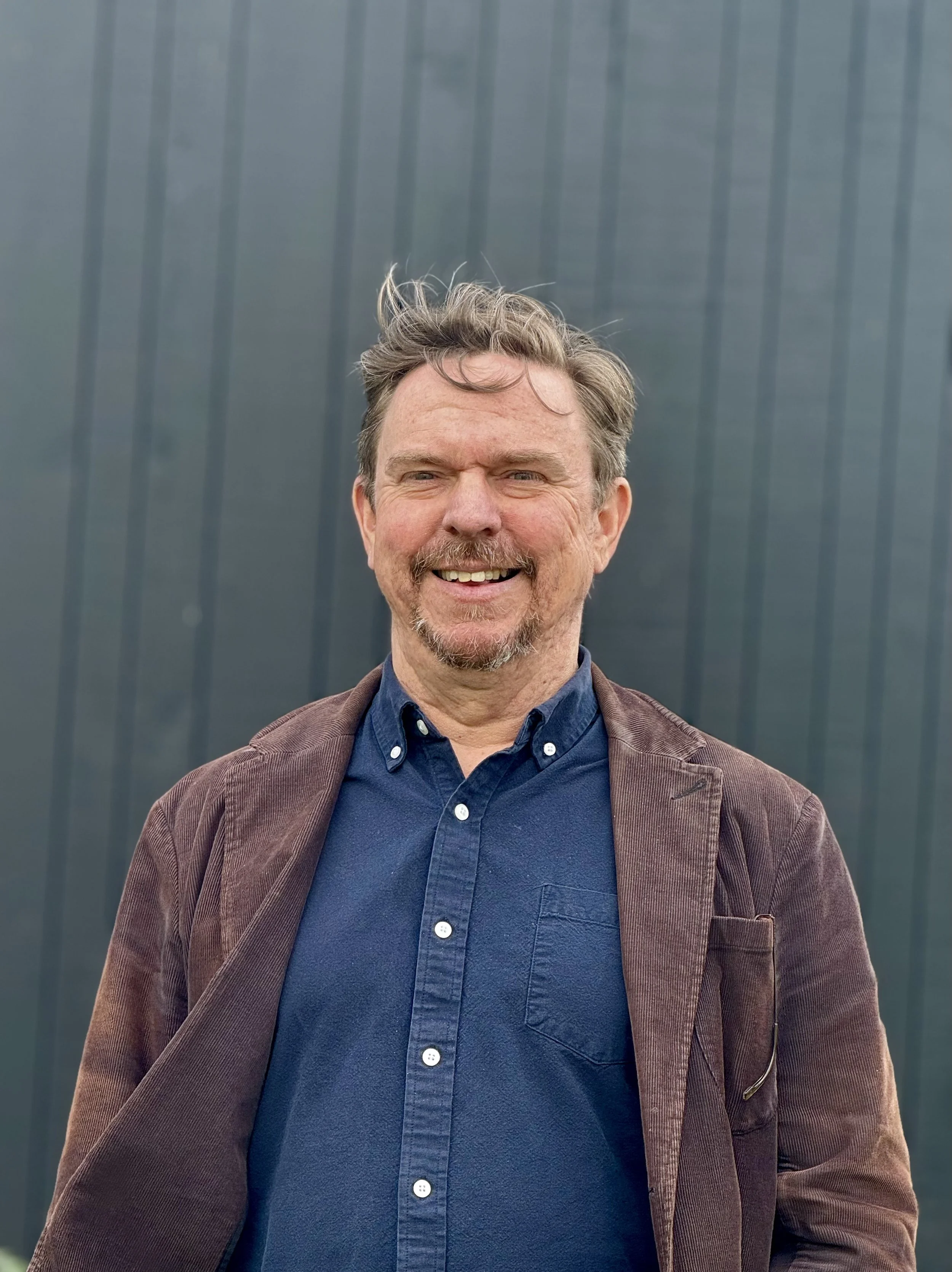 Portrait of a middle aged person with short brown hair and a goatee wearing a brown courderoy jacket and dark blue button down shirt in front of a dark gray background