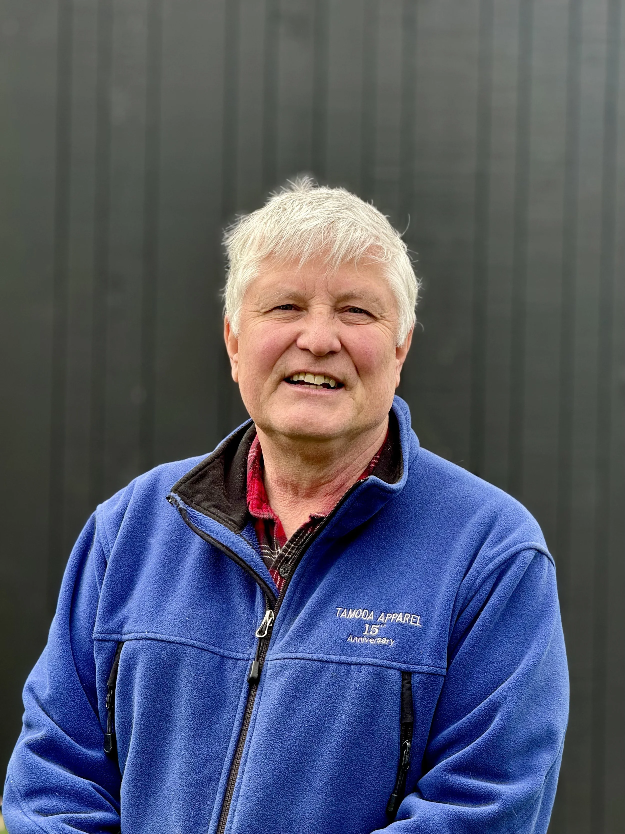 Portrait of an older person with short gray straight hair in blue fleece jacket in front of dark gray background